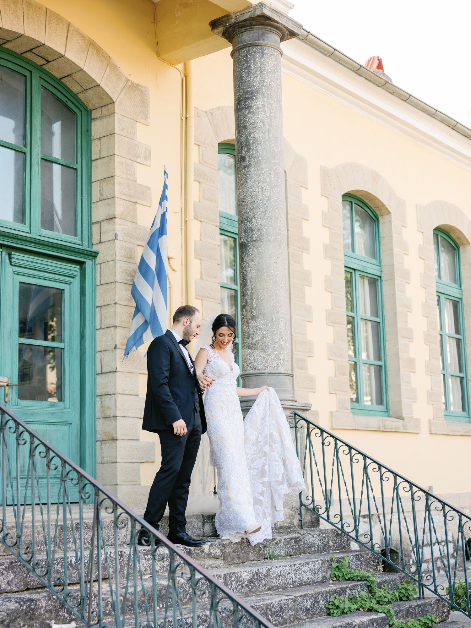 Newlyweds stand on stone steps of historic building with Greek flag.