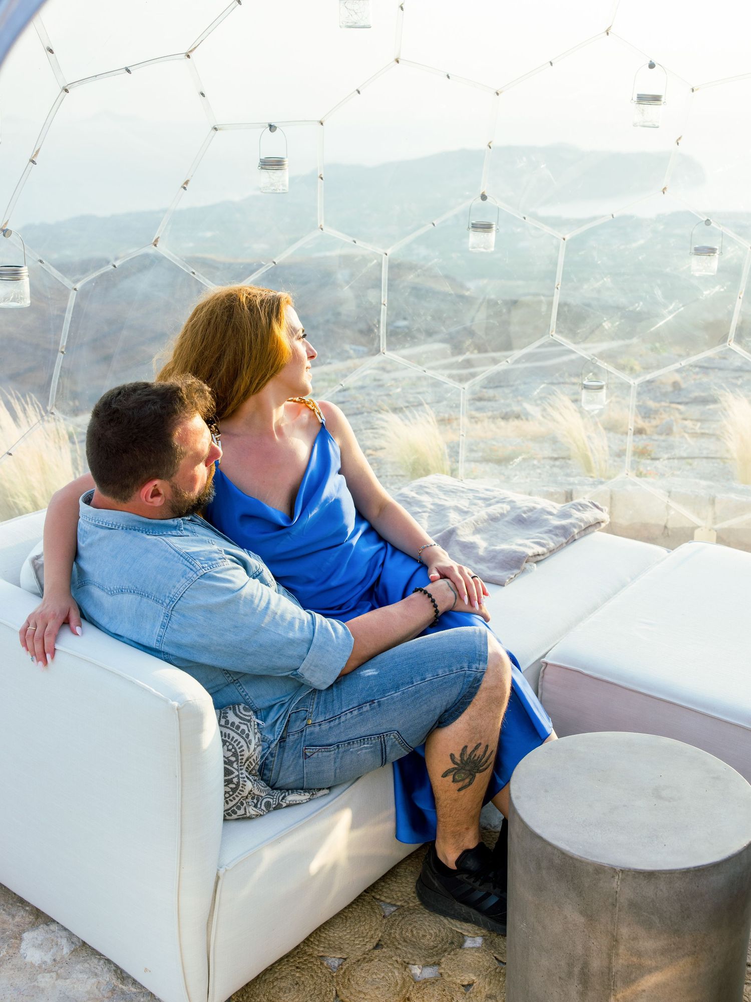 Couple relaxing on white outdoor sofa with geometric dome and mountain views.