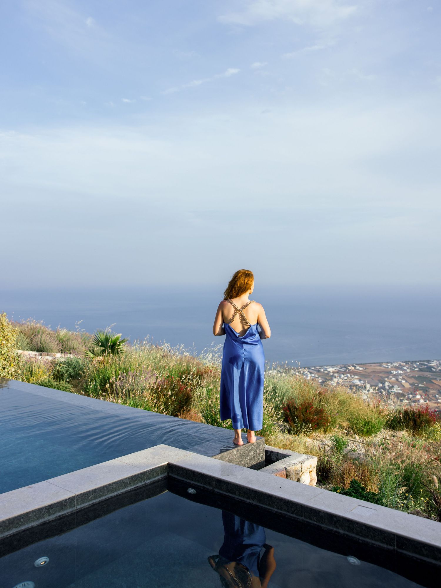Woman in blue dress standing at edge of infinity pool overlooking coastal views.