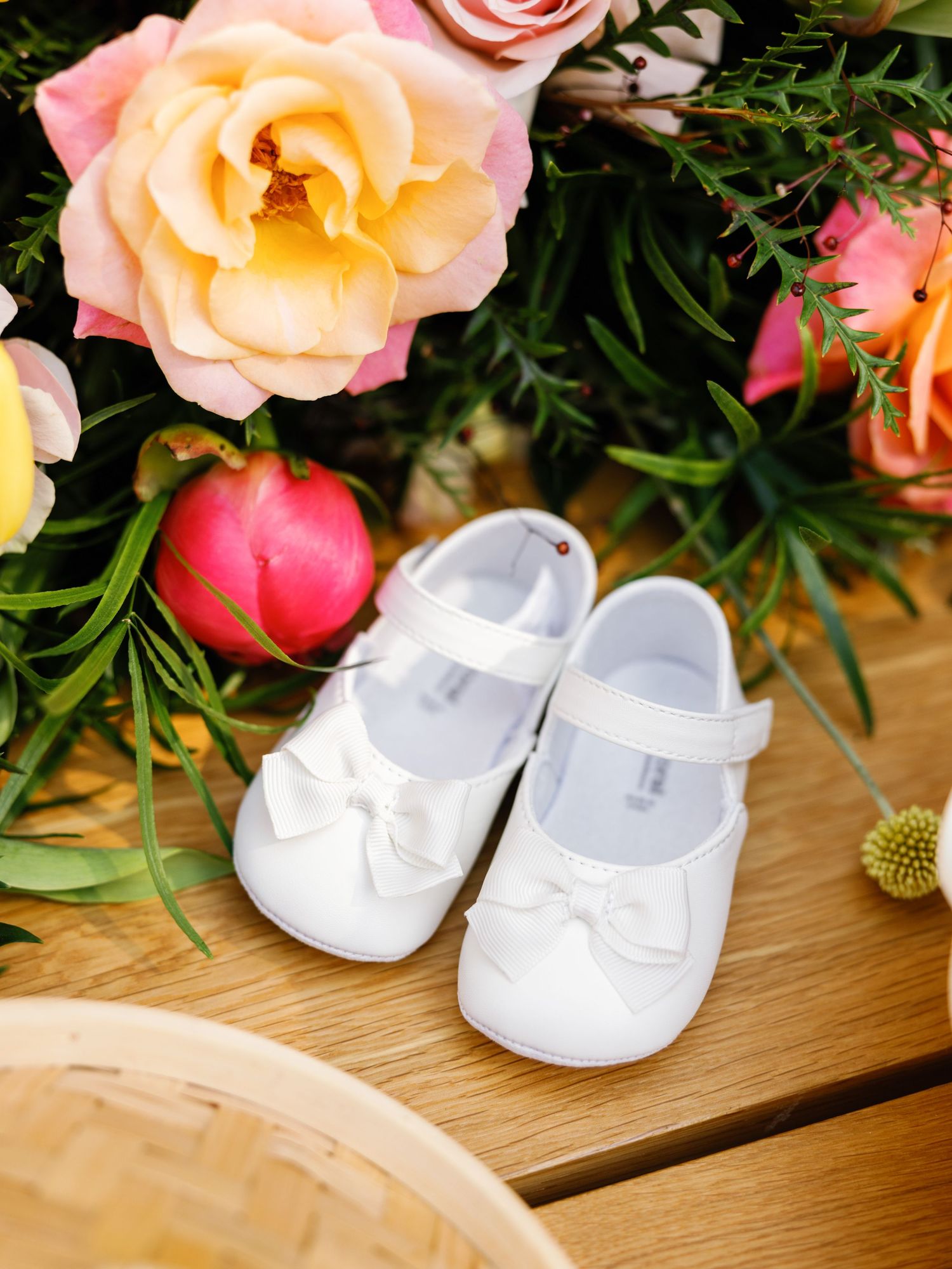 A pair of small white baby shoes with bows displayed next to pink and peach roses on wooden planks.