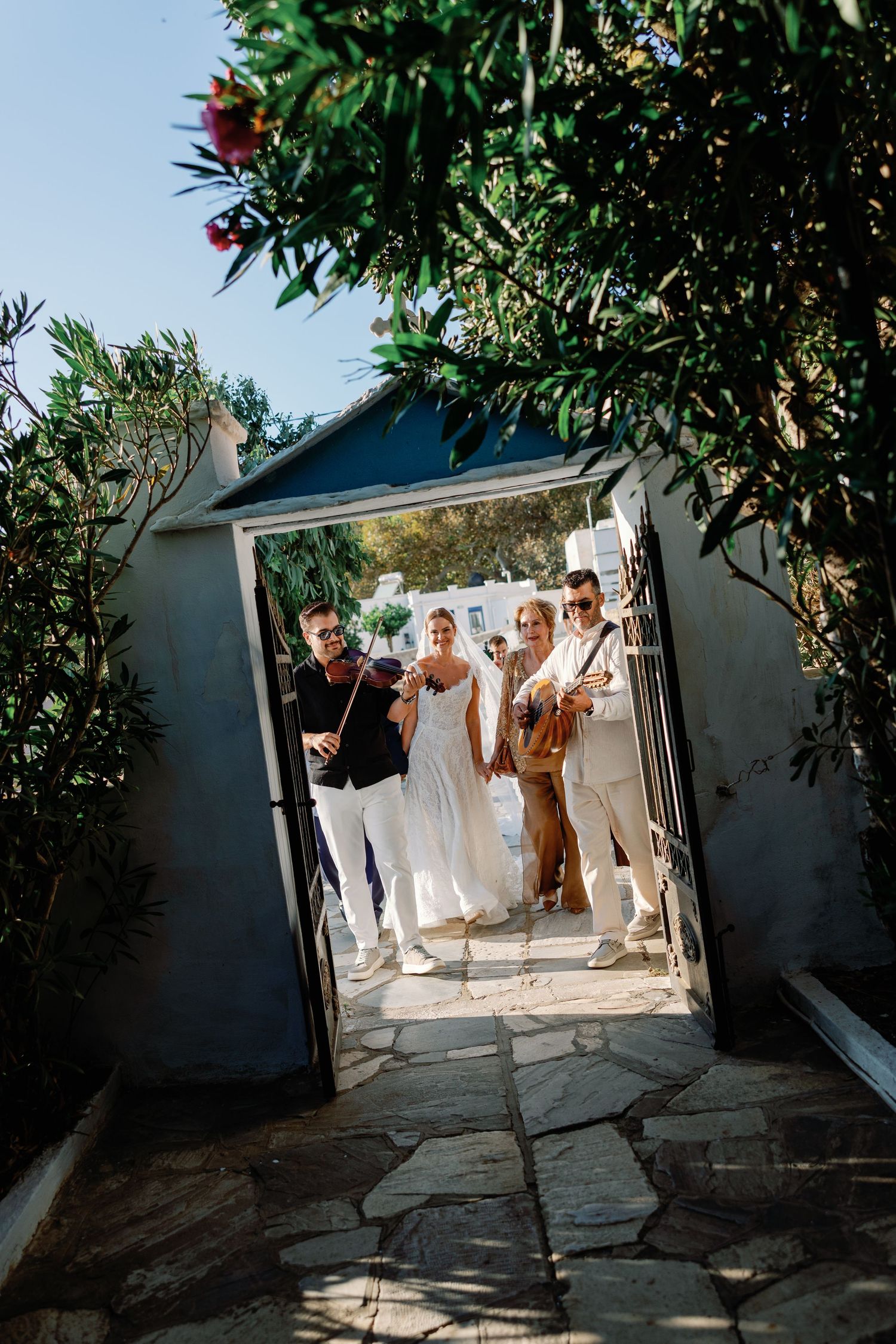 Wedding party entering through a stone archway gate, with musicians playing violin and guitar outdoors.