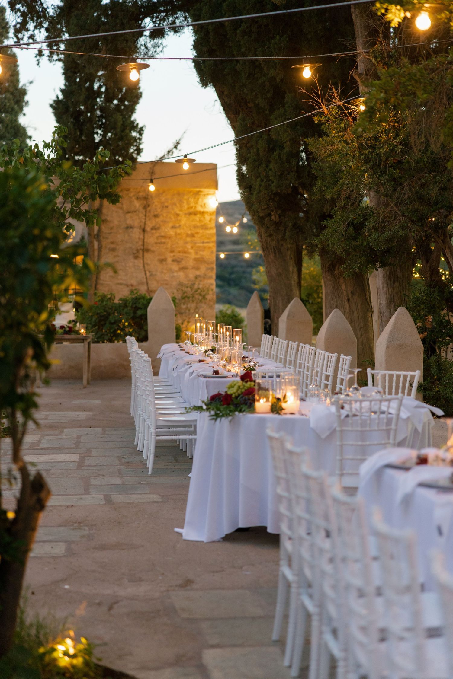 Elegant outdoor wedding reception with white Chiavari chairs, long tables, floral centerpieces, and string lights at dusk.