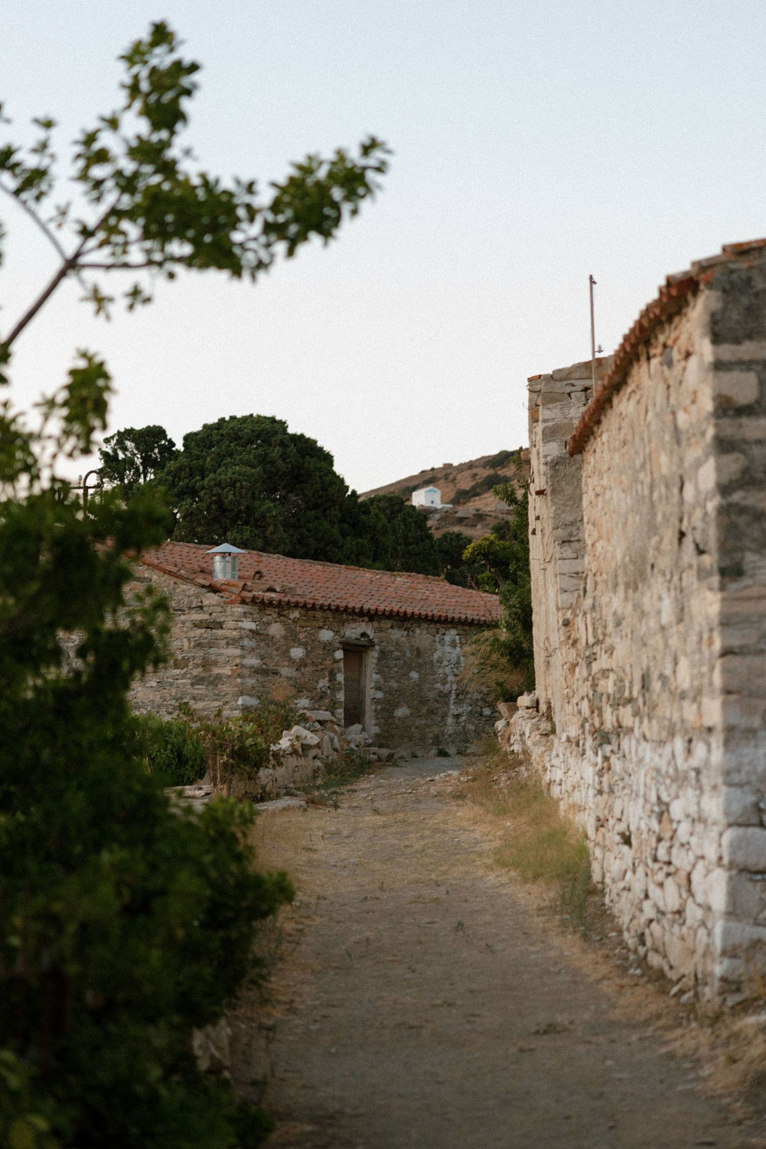 A narrow stone path winds between ancient stone walls leading to a rustic building with red tile roof in a Mediterranean village.