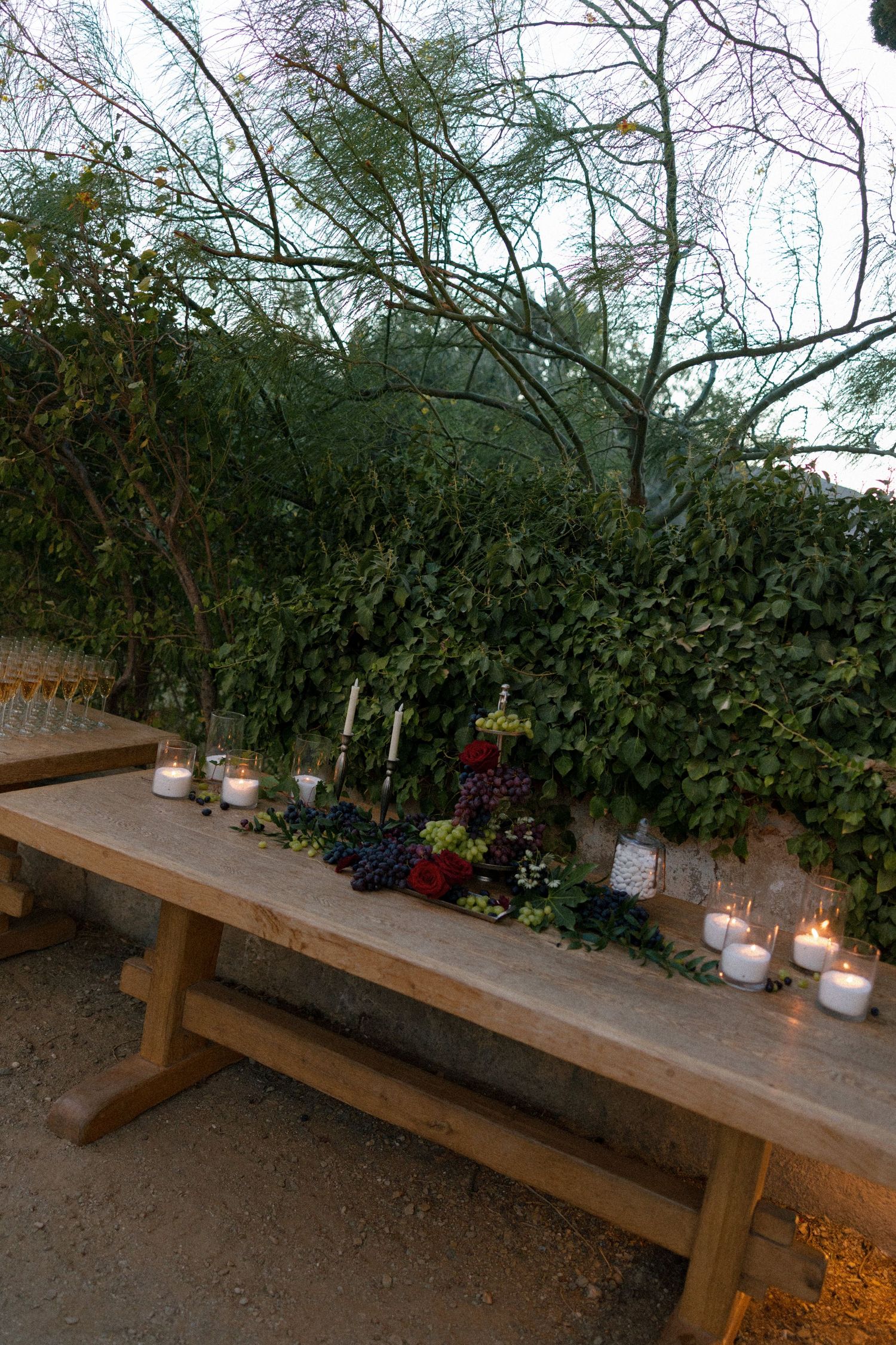 Rustic wooden outdoor banquet table decorated with candles, grapes, and greenery against a lush ivy wall at dusk.