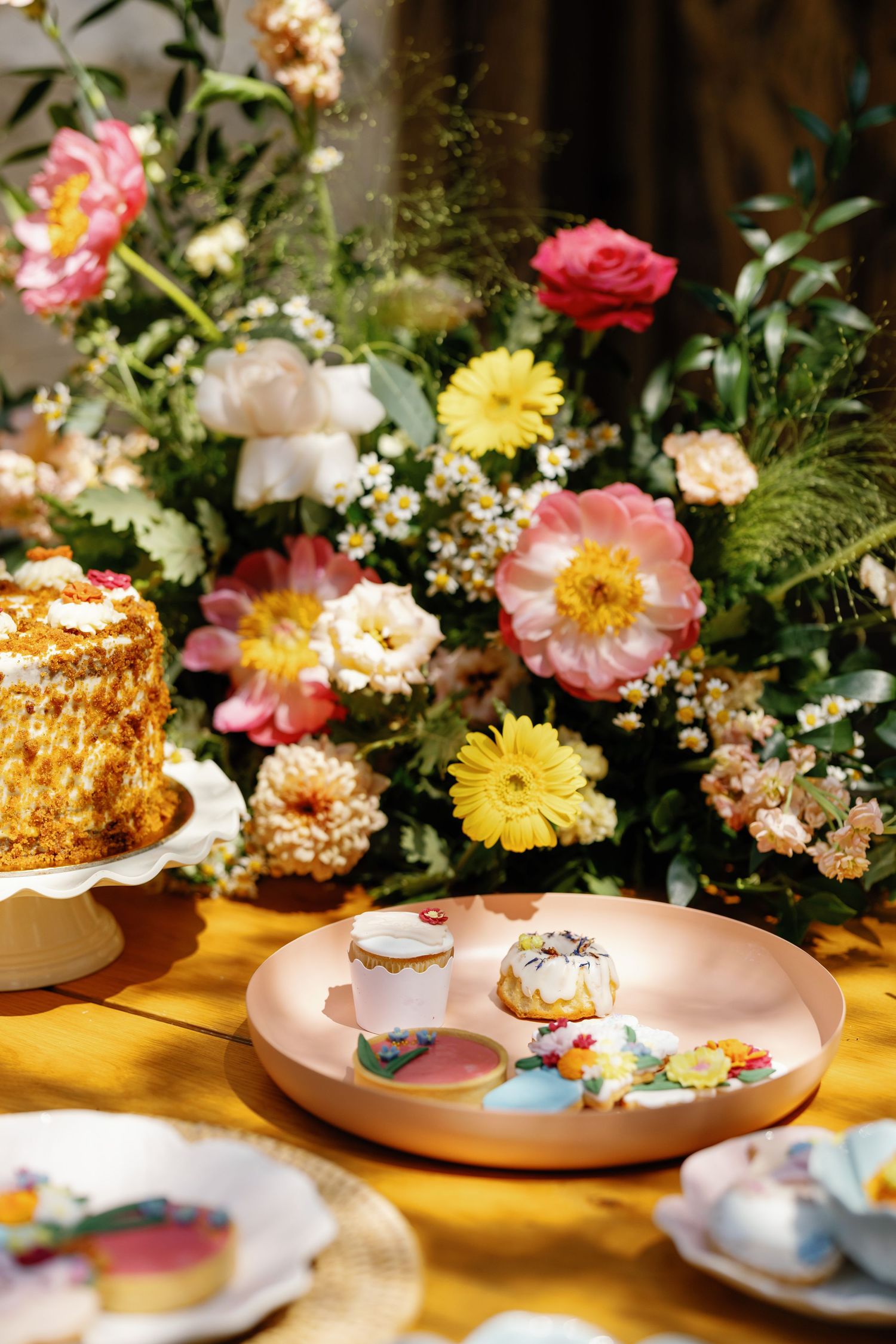 Elegant dessert display with colorful floral arrangement featuring roses, gerberas and a layered cake on a wooden table.