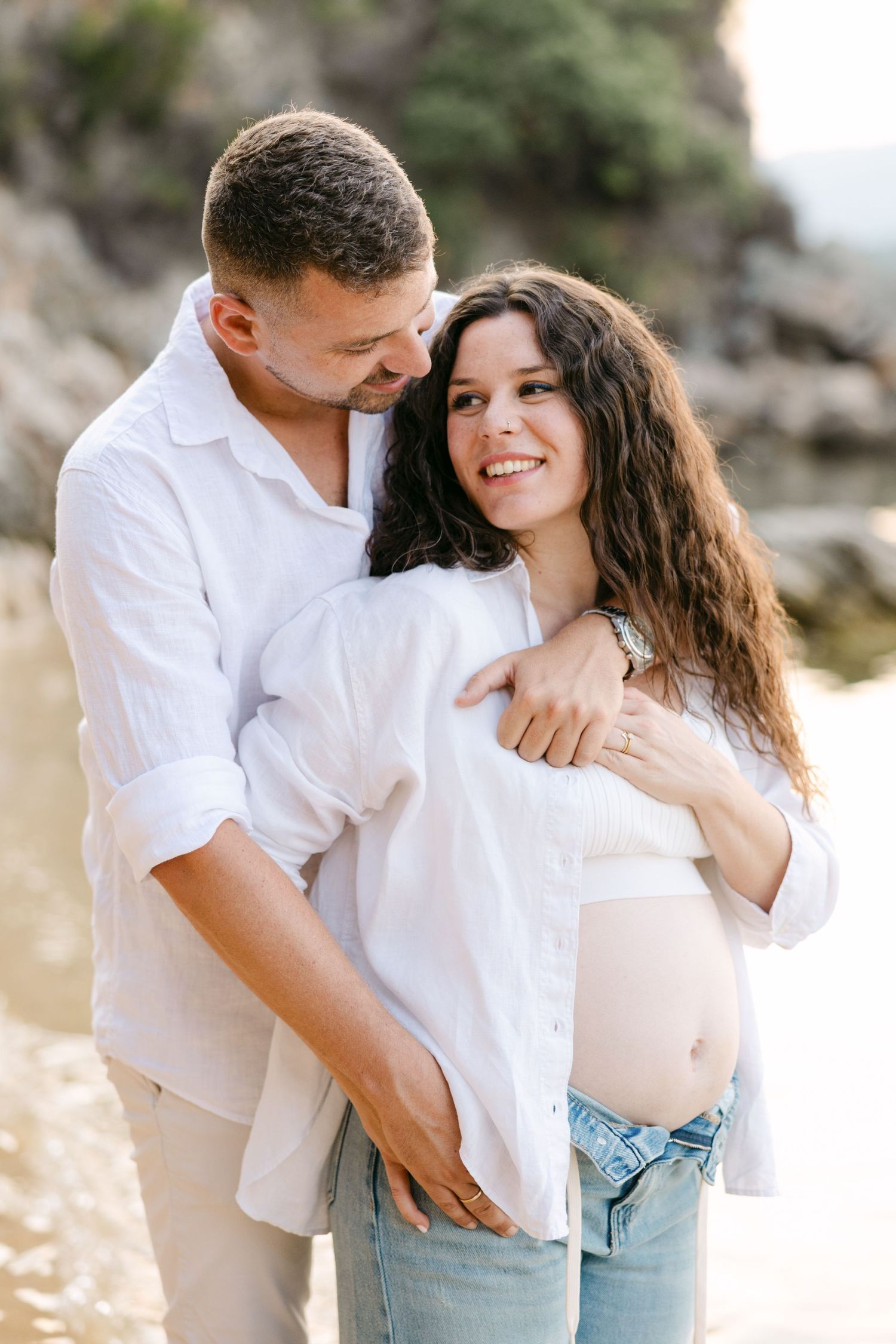 An intimate maternity portrait featuring a couple in matching white shirts against a natural outdoor backdrop.