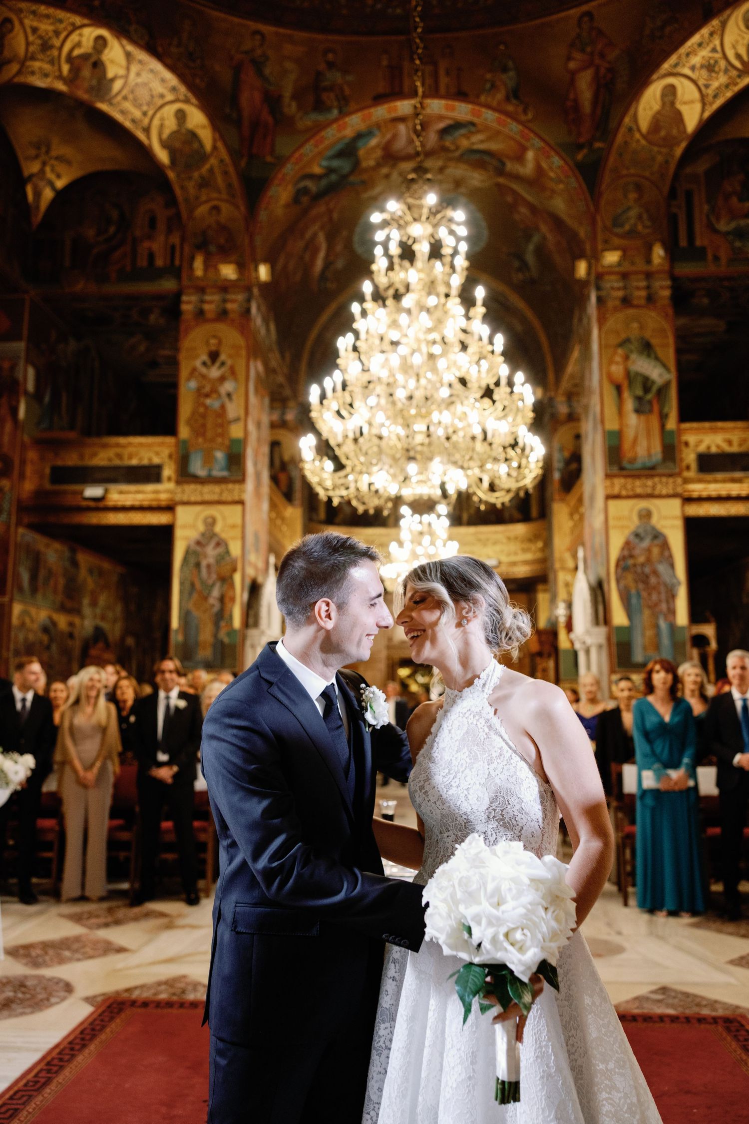 Elegant wedding couple sharing intimate moment in ornate church interior with crystal chandeliers.