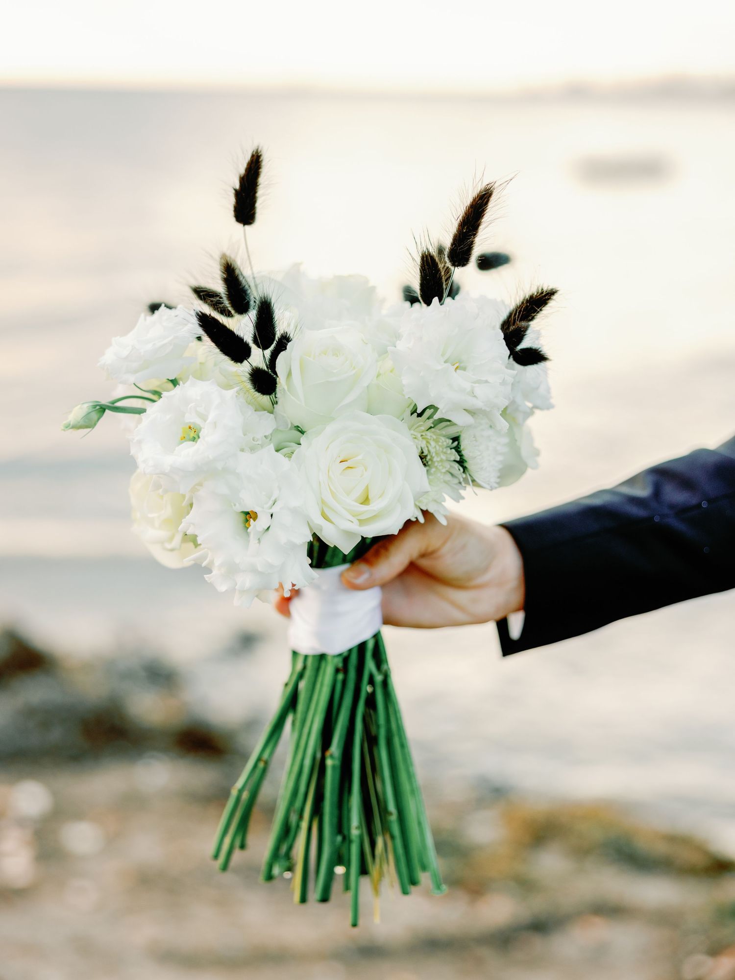 An elegant white rose wedding bouquet with dark feather accents held against a sunset backdrop.