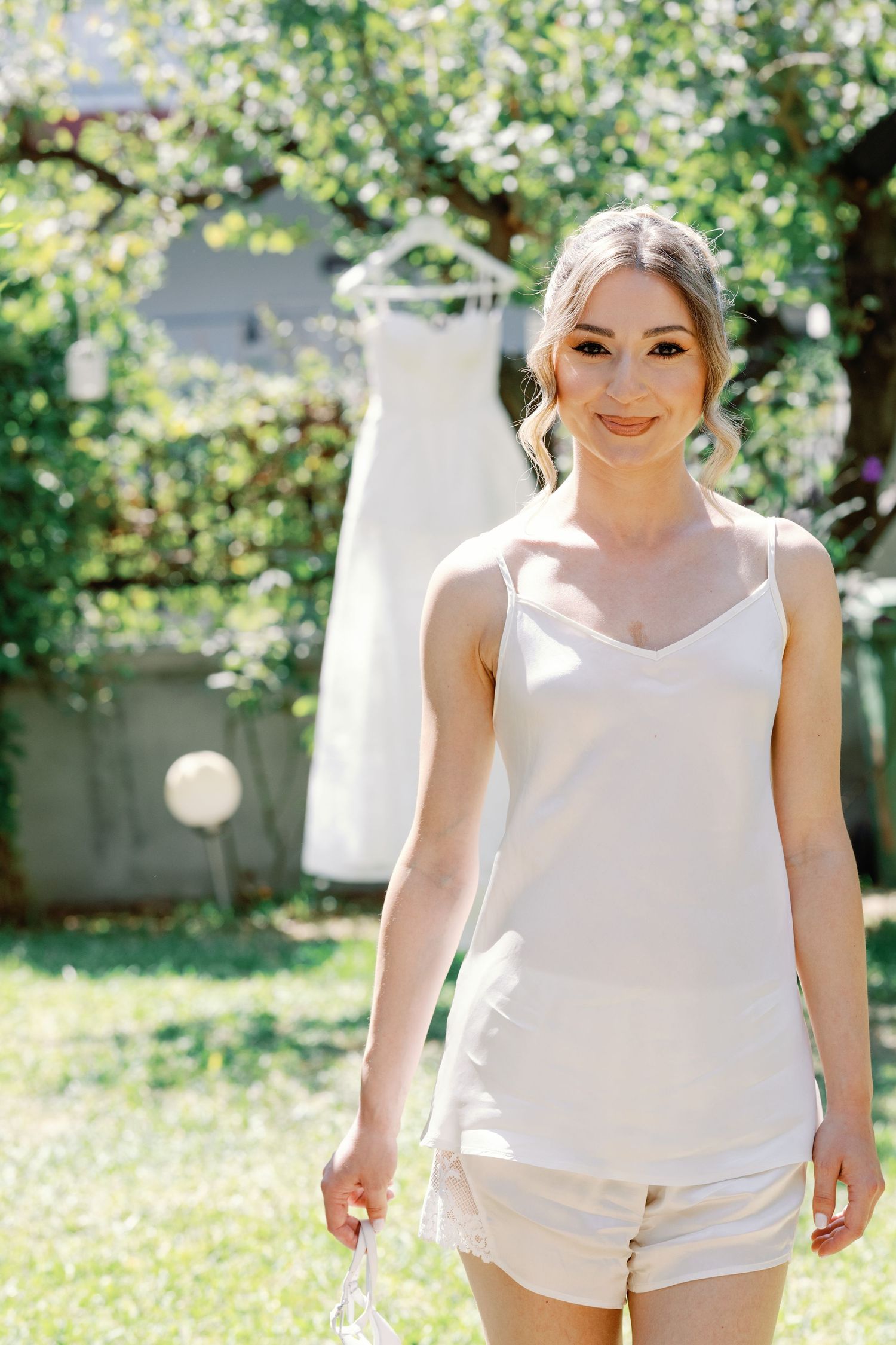 A person in white clothing poses outdoors in a sunny garden setting.