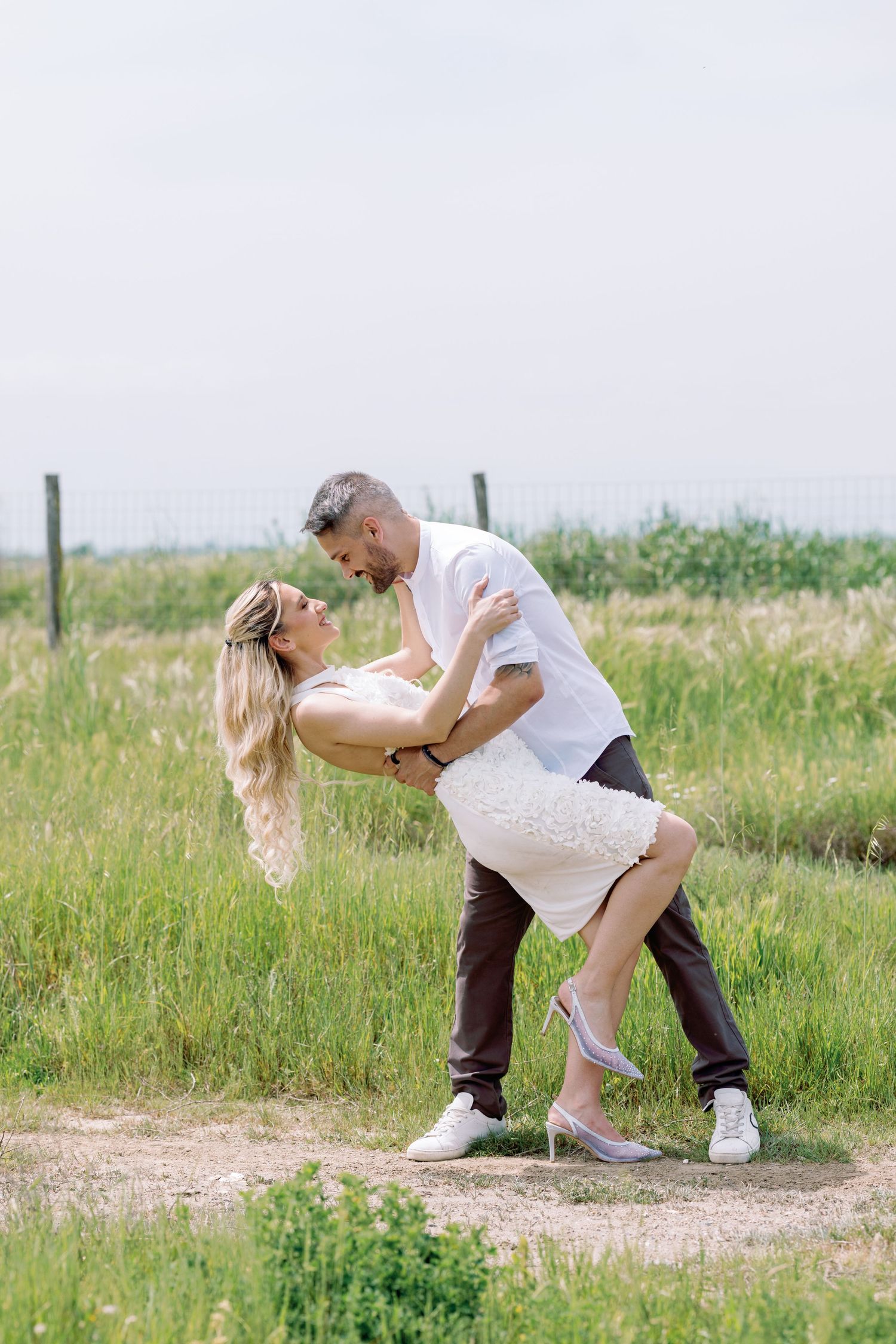 Couple dancing outdoors in a green field, man dipping woman in white dress and heels on a dirt path.