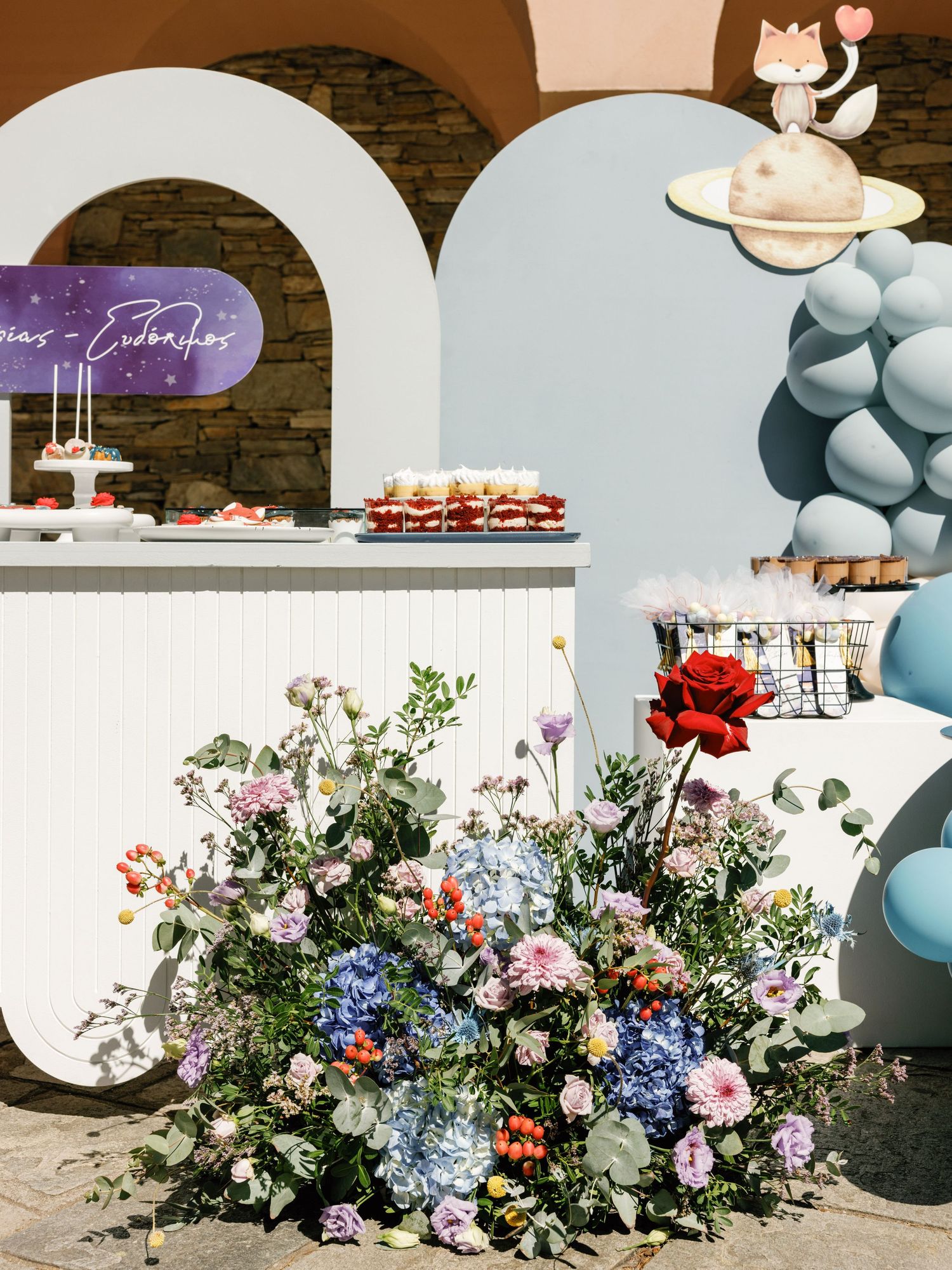 Elegant wedding dessert display with balloon arch and lush floral arrangements in blue, pink and white tones.