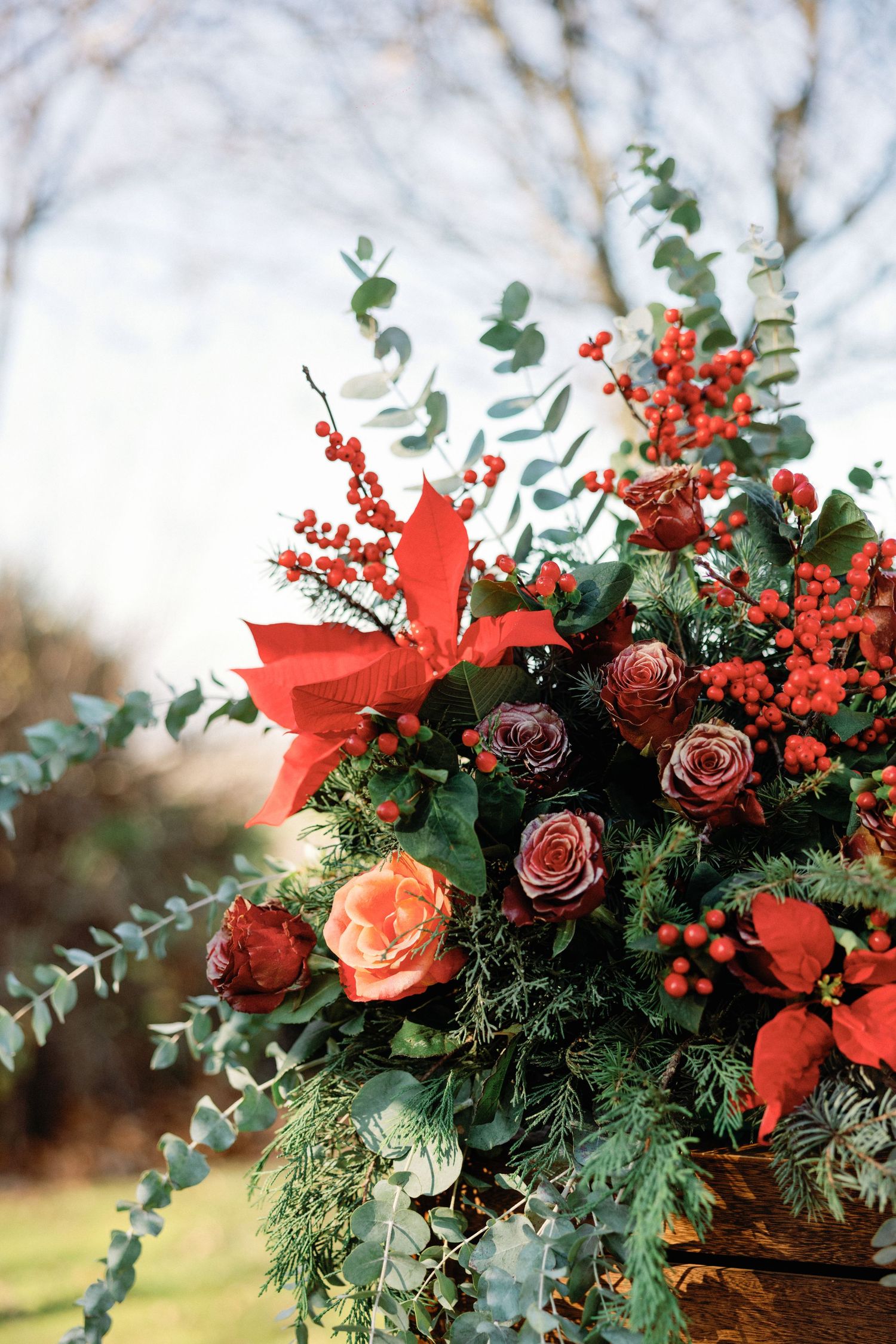 A festive holiday floral arrangement with red poinsettias, roses, berries, and eucalyptus greenery.