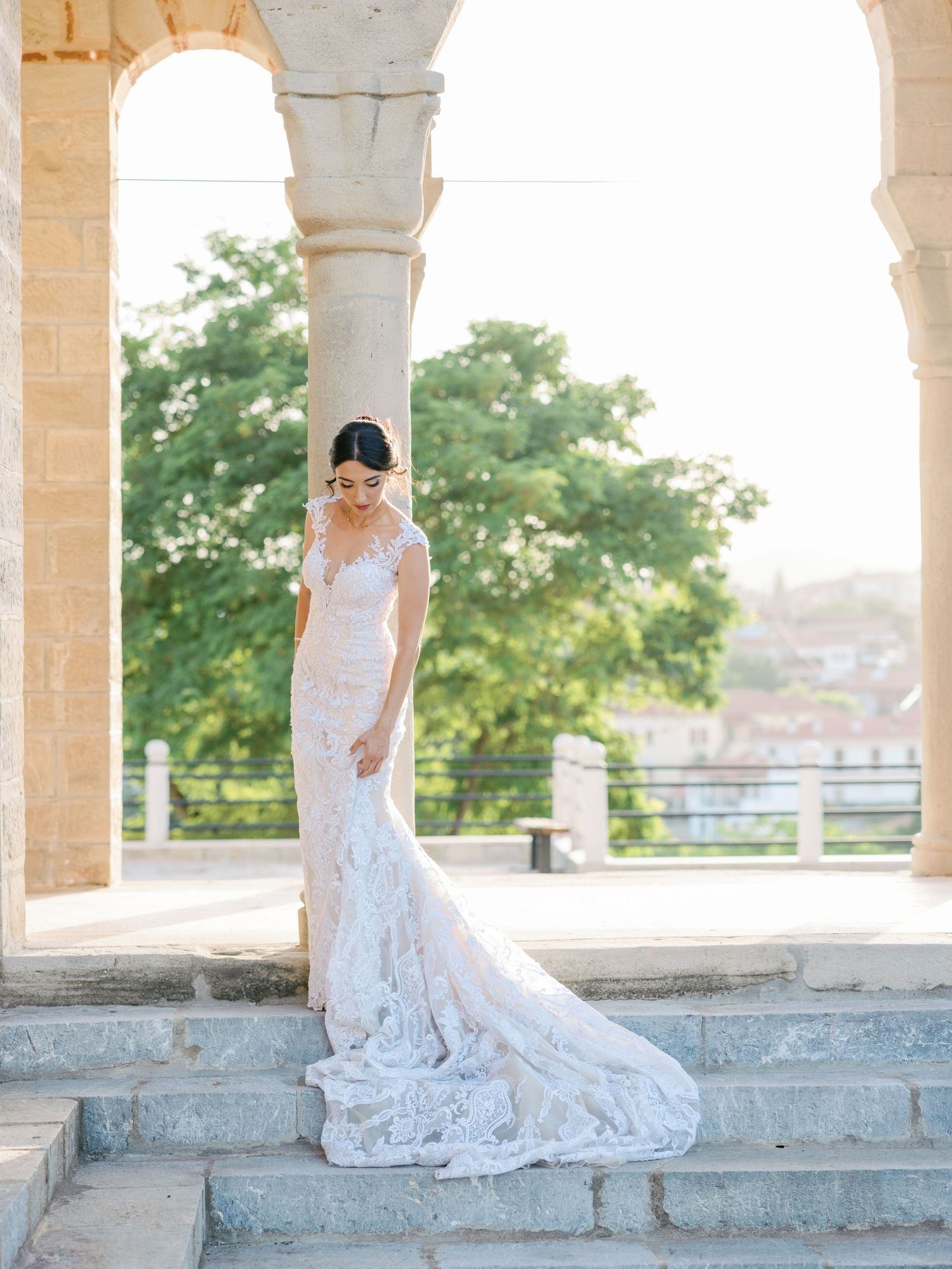 A bride in a fitted lace gown with flowing train stands gracefully on stone steps under arches.