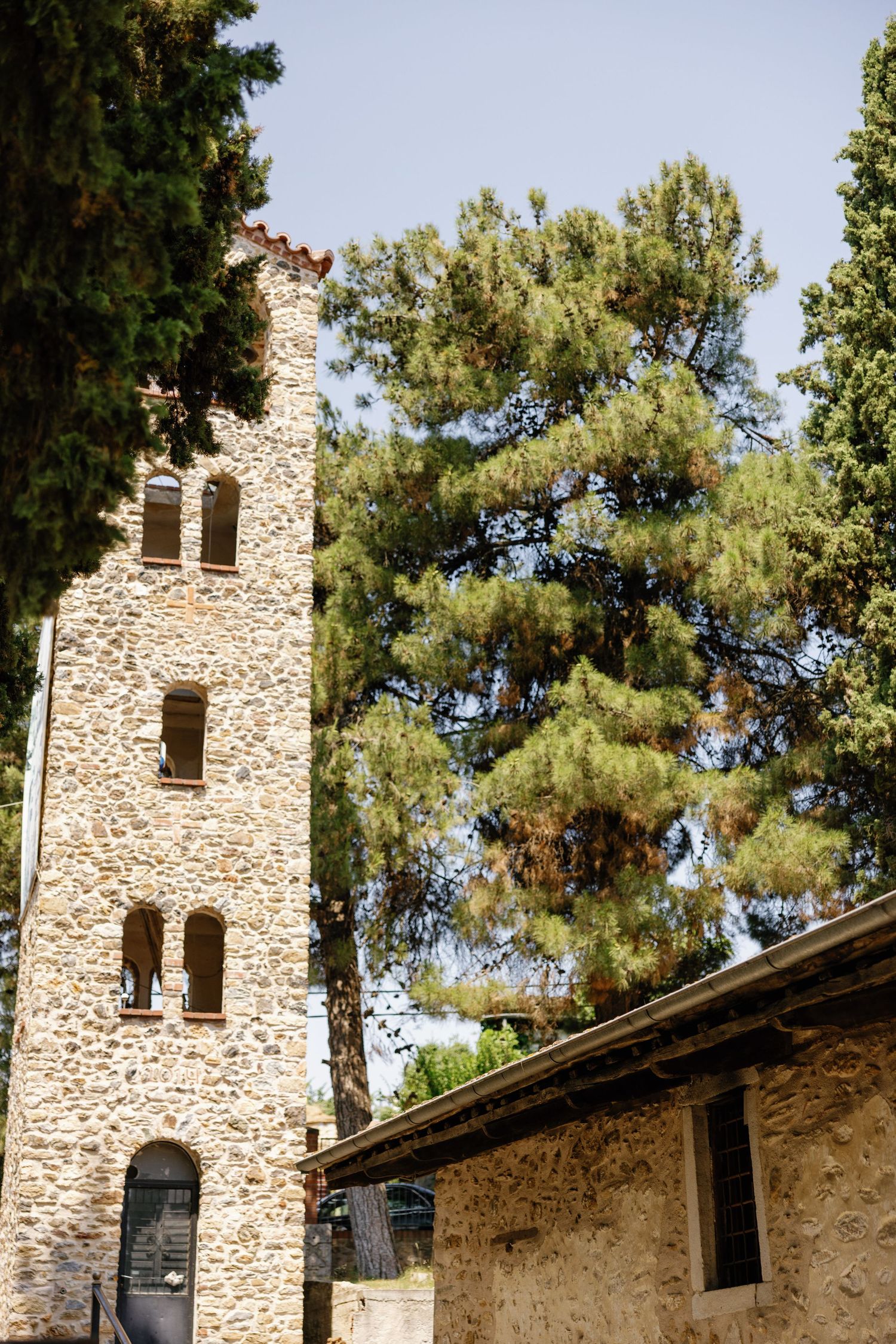 Ancient stone tower with arched windows surrounded by tall pine trees against blue sky.