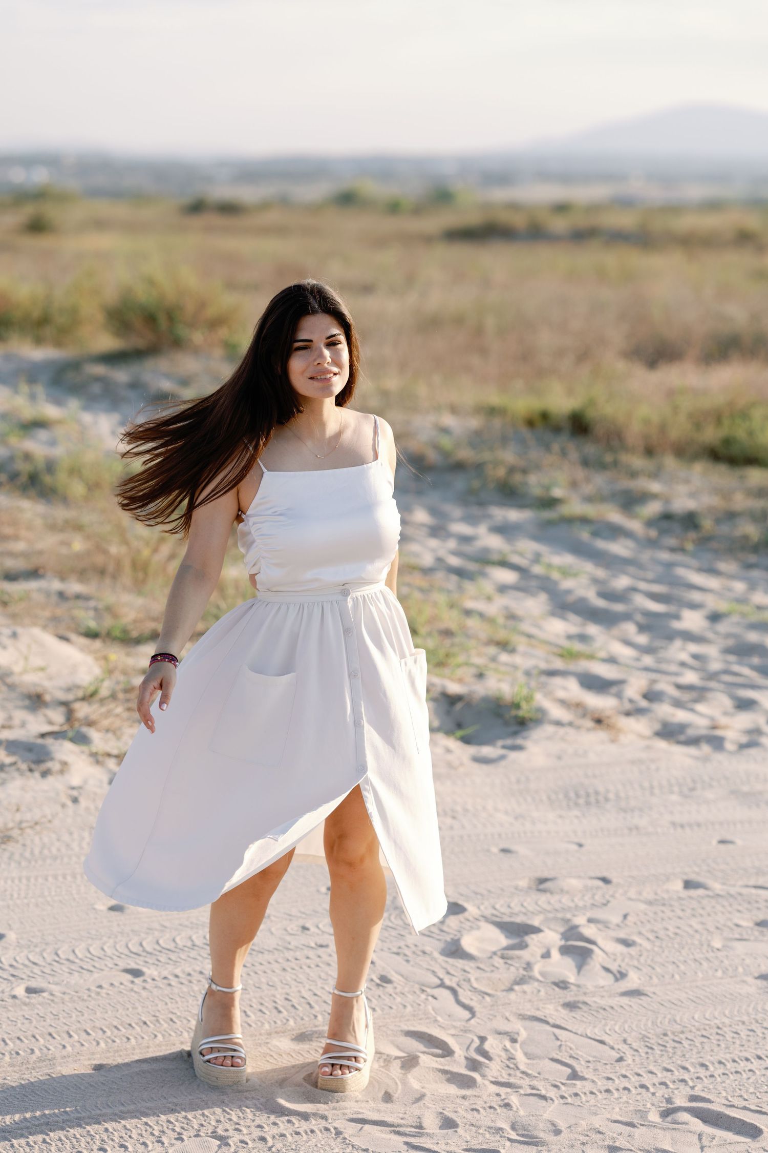 Woman in white dress twirling playfully on sandy beach during photoshoot.