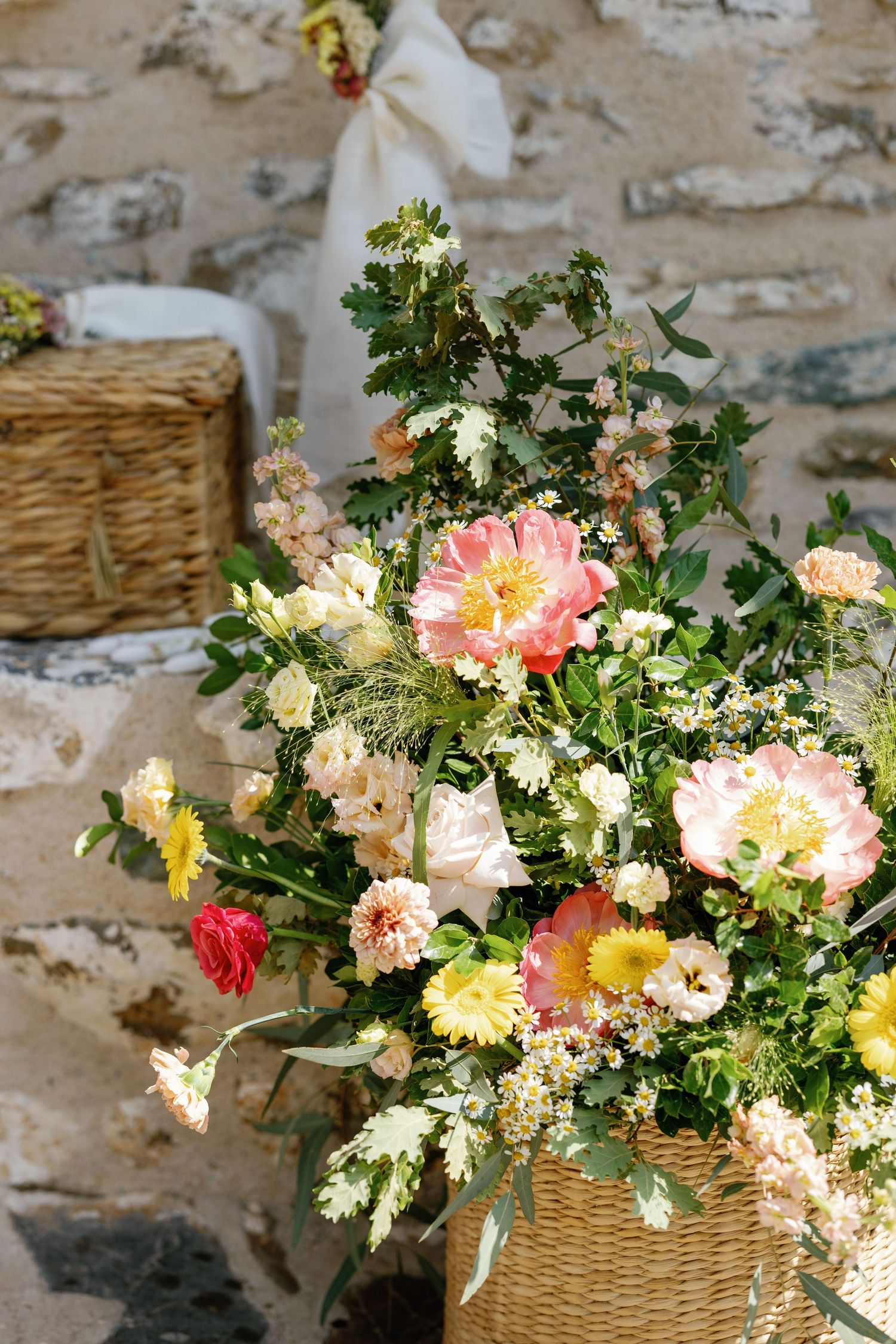 A lush floral arrangement with pink and yellow blooms in a woven basket against stone wall.