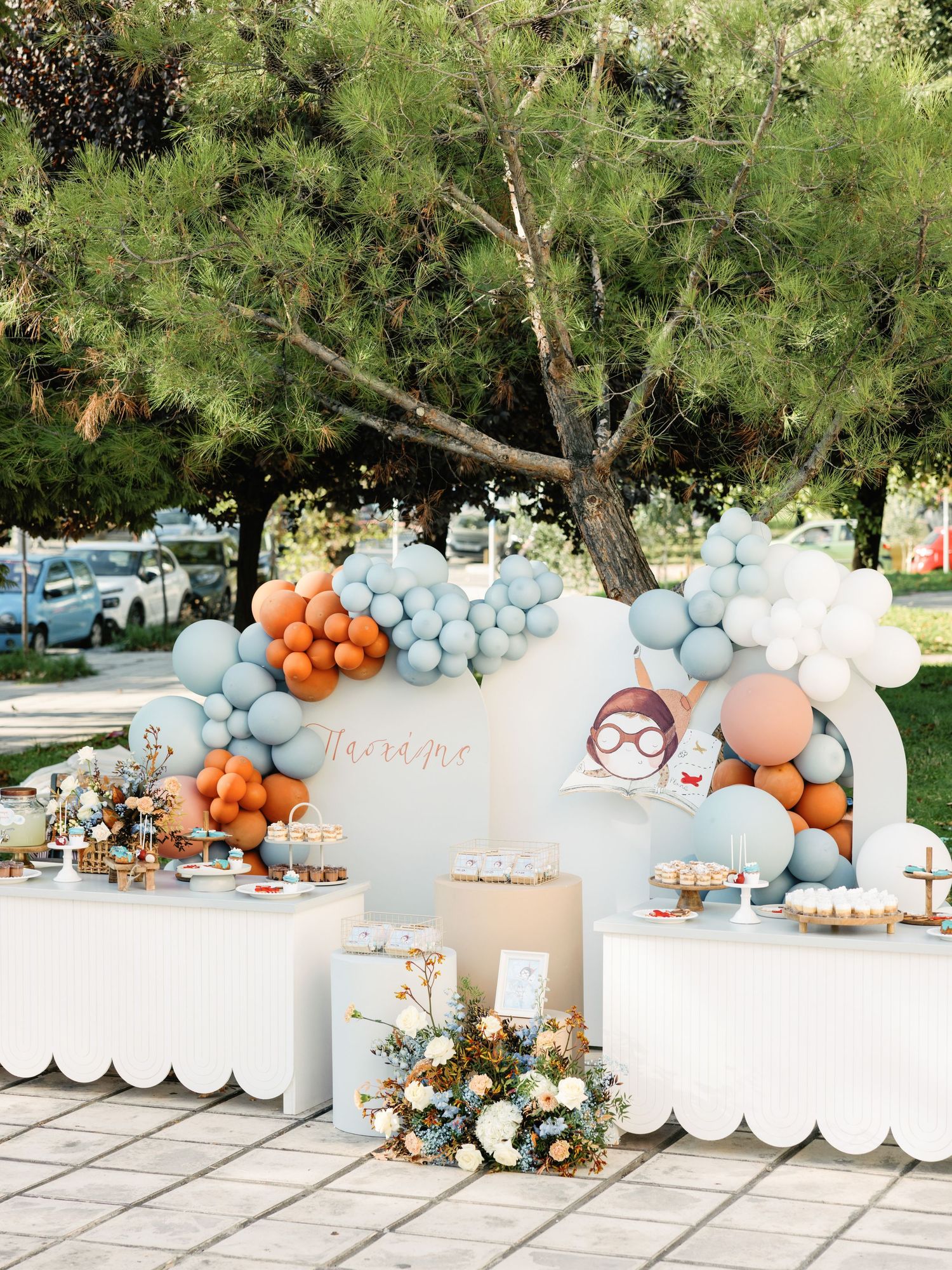 Elegant party setup with white and peach balloon arch, scalloped table, and floral arrangements under a pine tree.