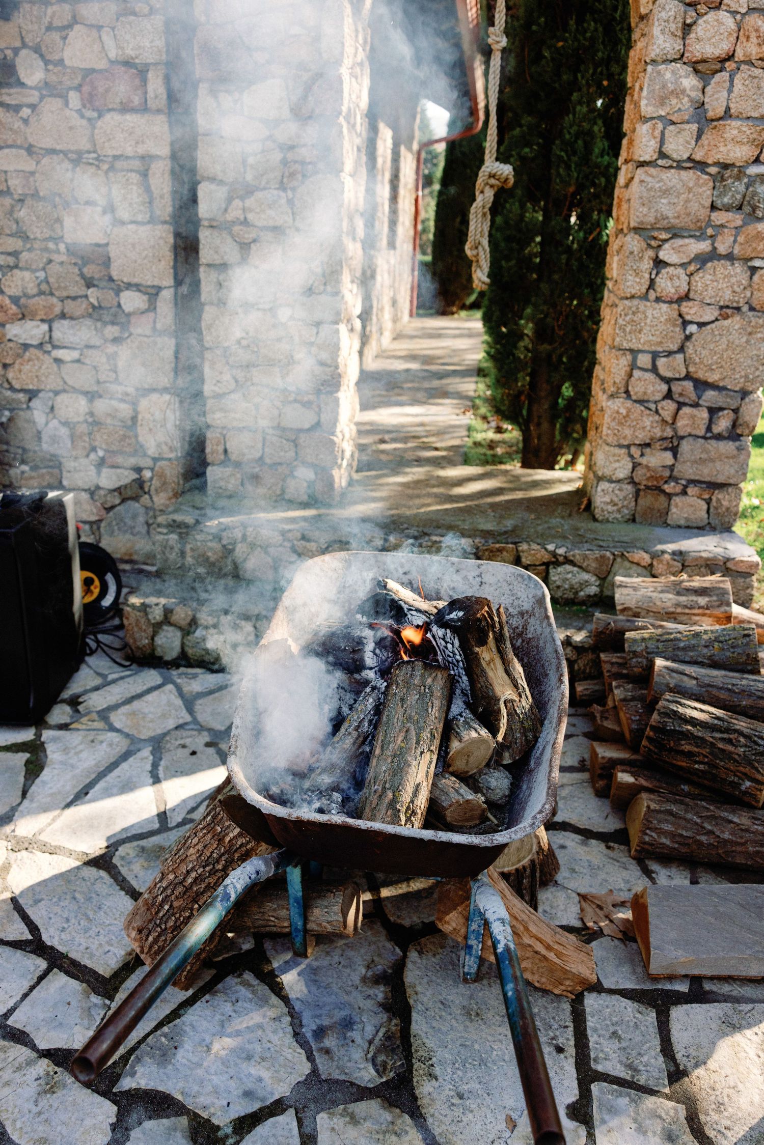 A rustic outdoor cooking setup with burning firewood in a stone-walled courtyard creating atmospheric smoke.