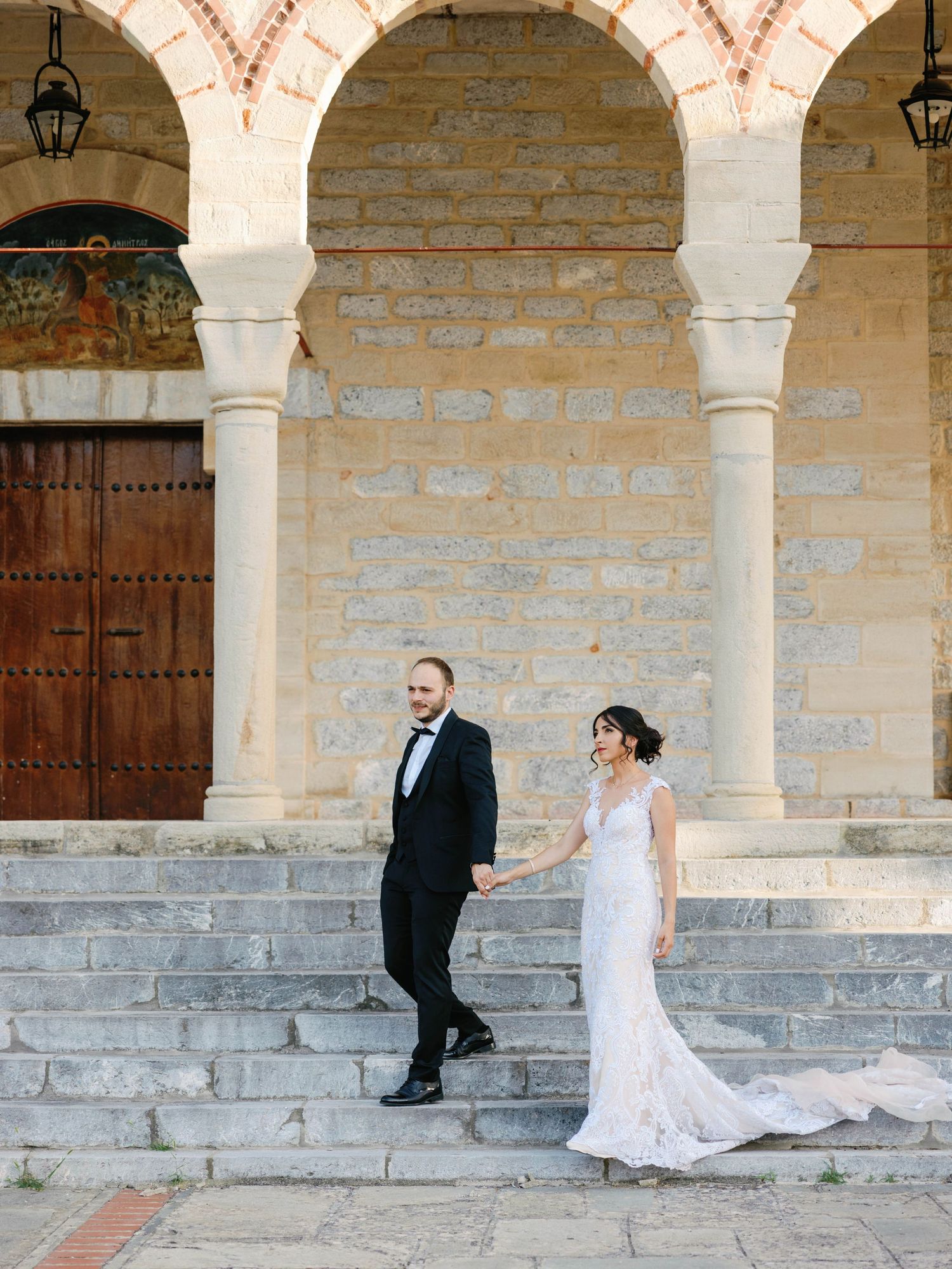 A couple in wedding attire poses elegantly on stone steps beneath ornate archways of a historic building.