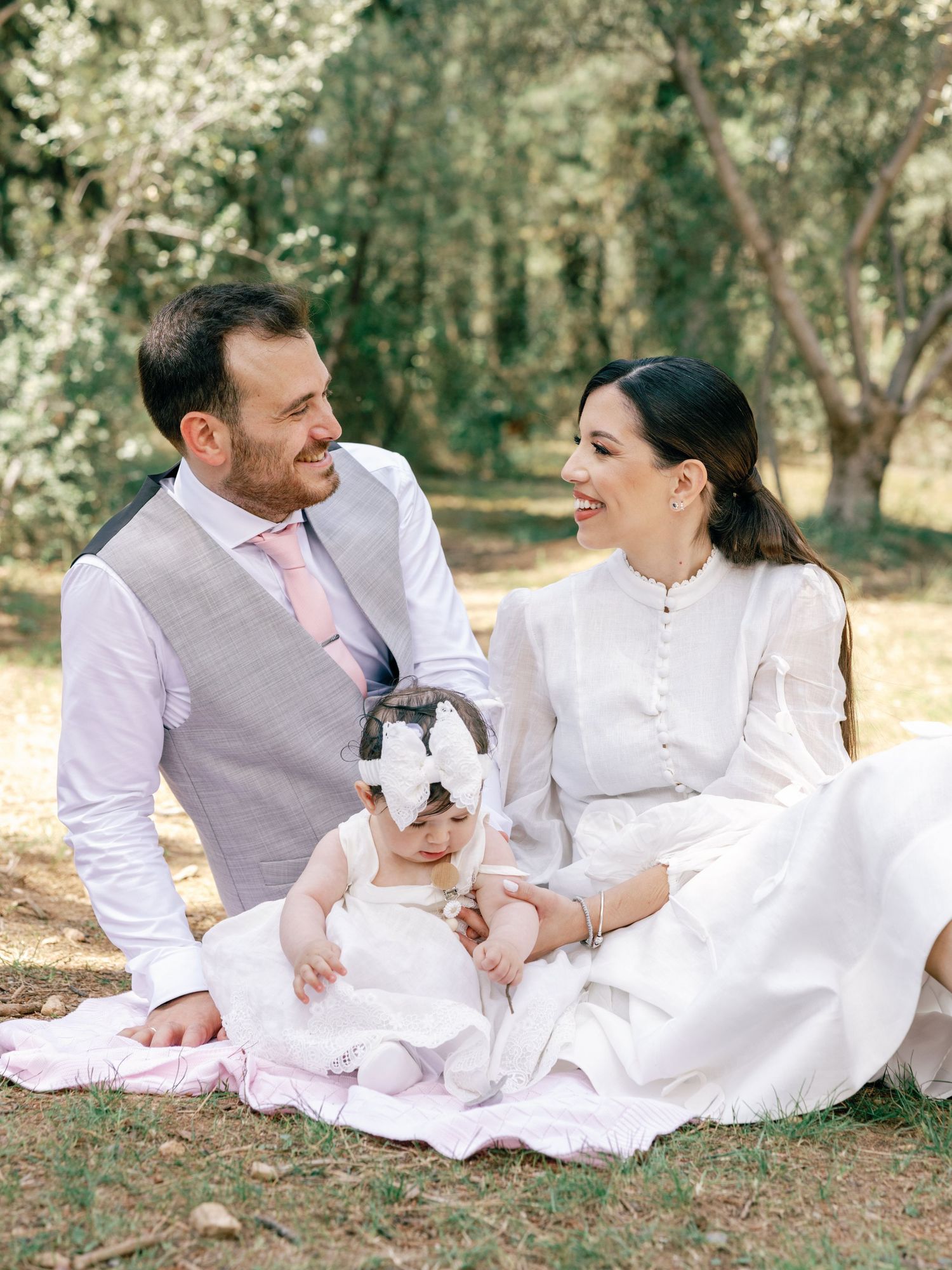 A couple in white attire shares intimate moments while relaxing on a blanket in a sun-dappled outdoor setting.