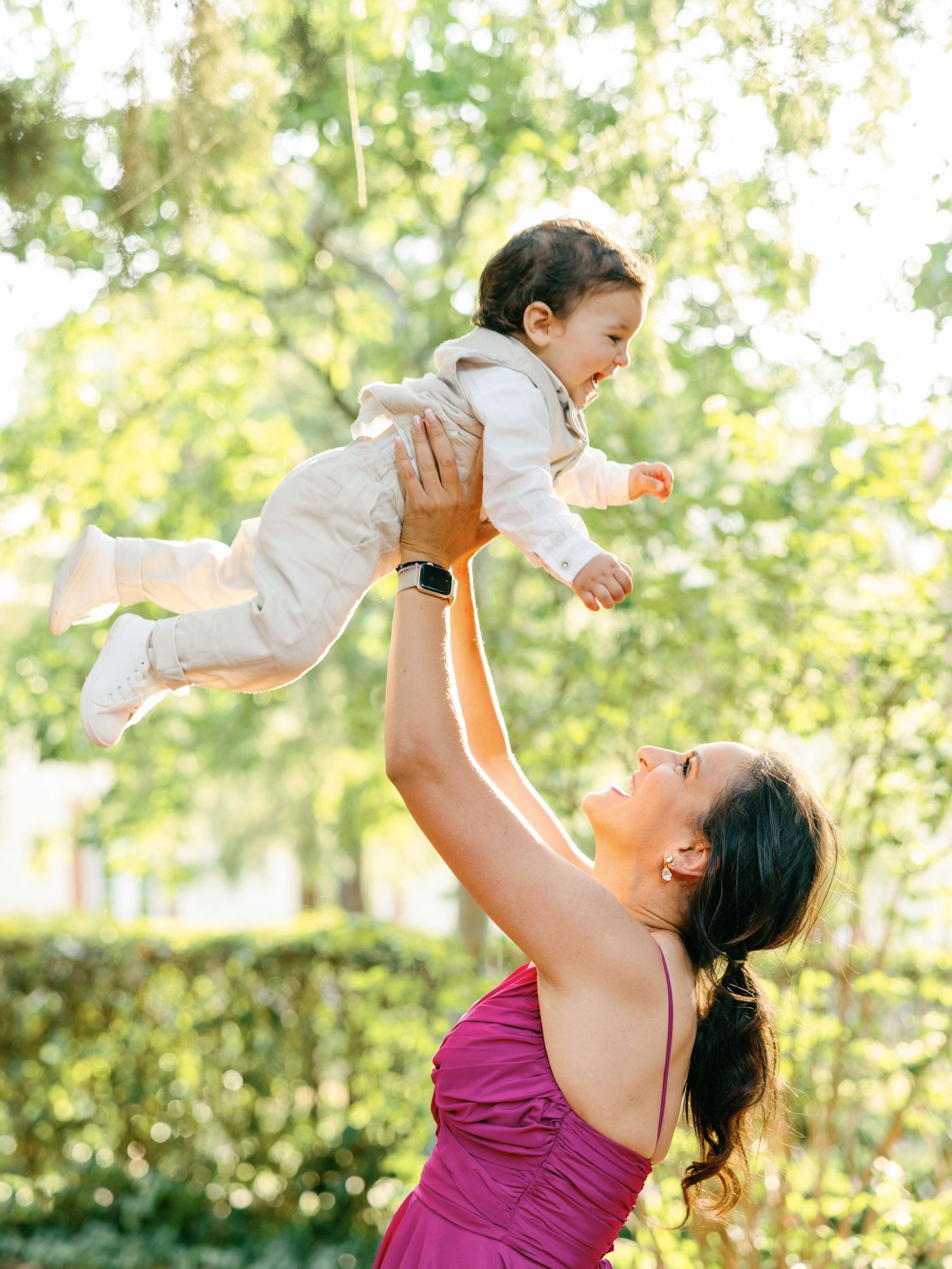 A joyful moment captured outdoors as a baby in white is lifted high into the air against a sunlit garden background.