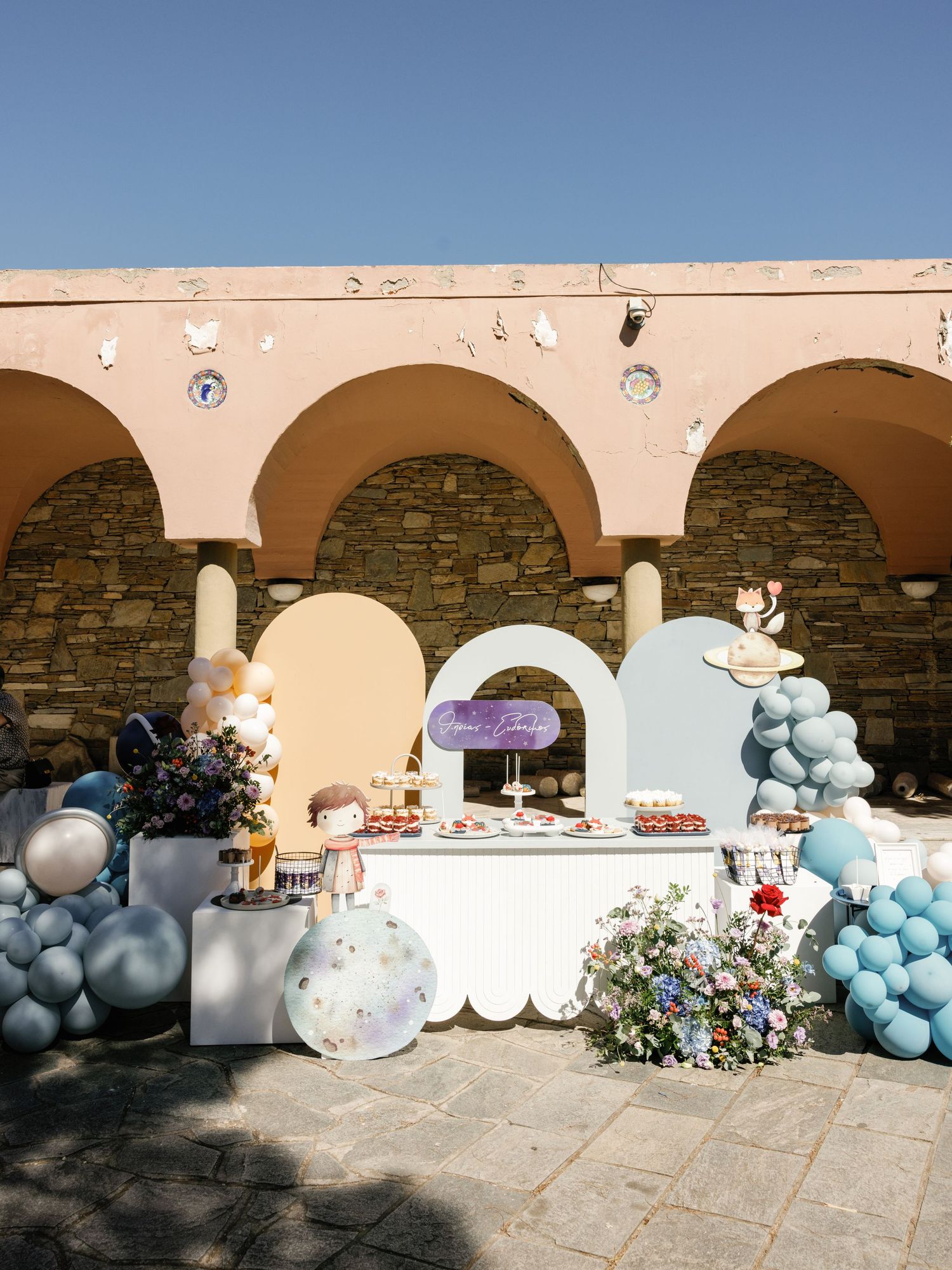 Decorative balloon arch installation with blue and white balloons adorning an outdoor stone archway against a sunny sky.