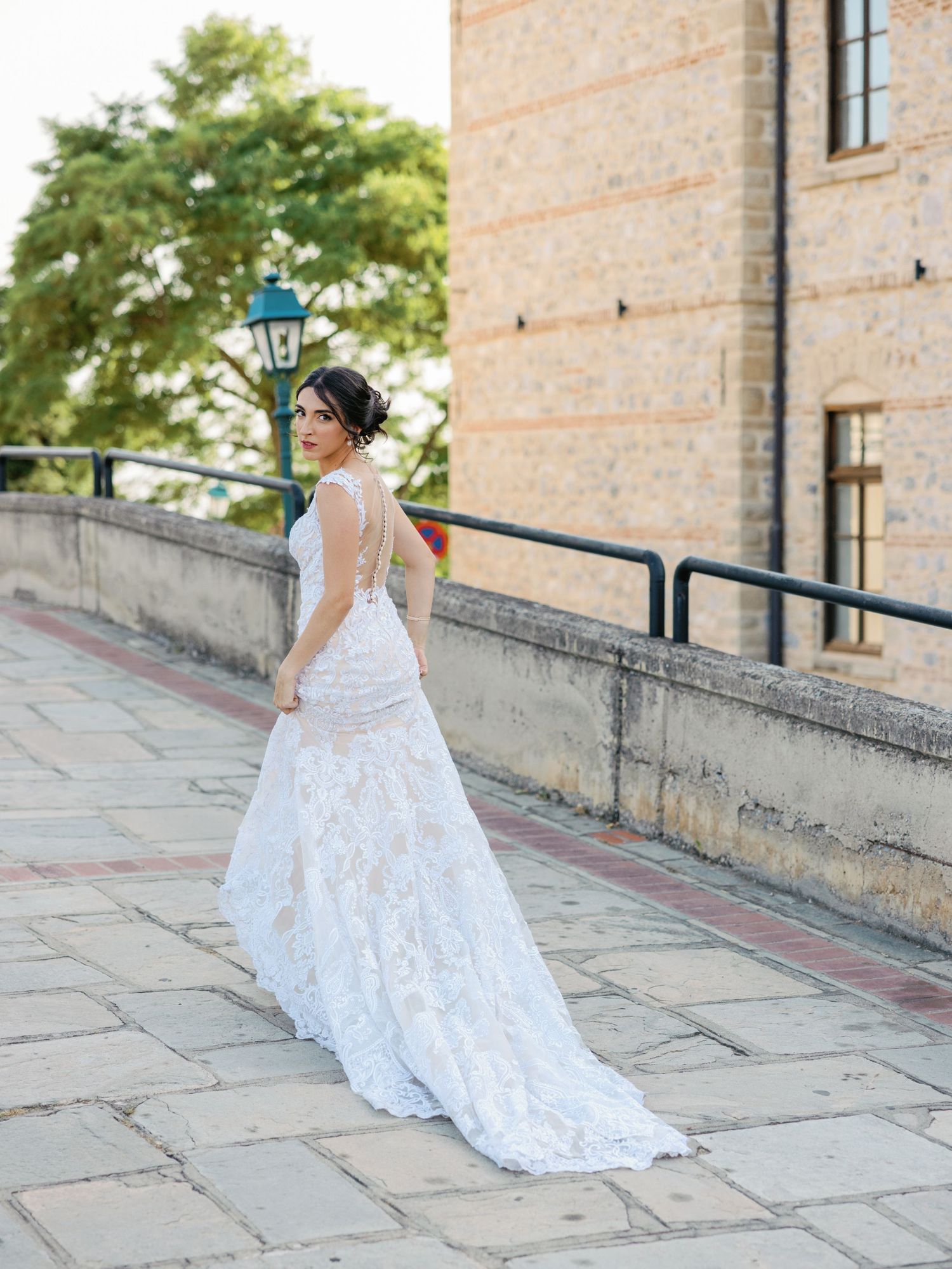Flowing white lace wedding dress trailing on stone path near historic building.