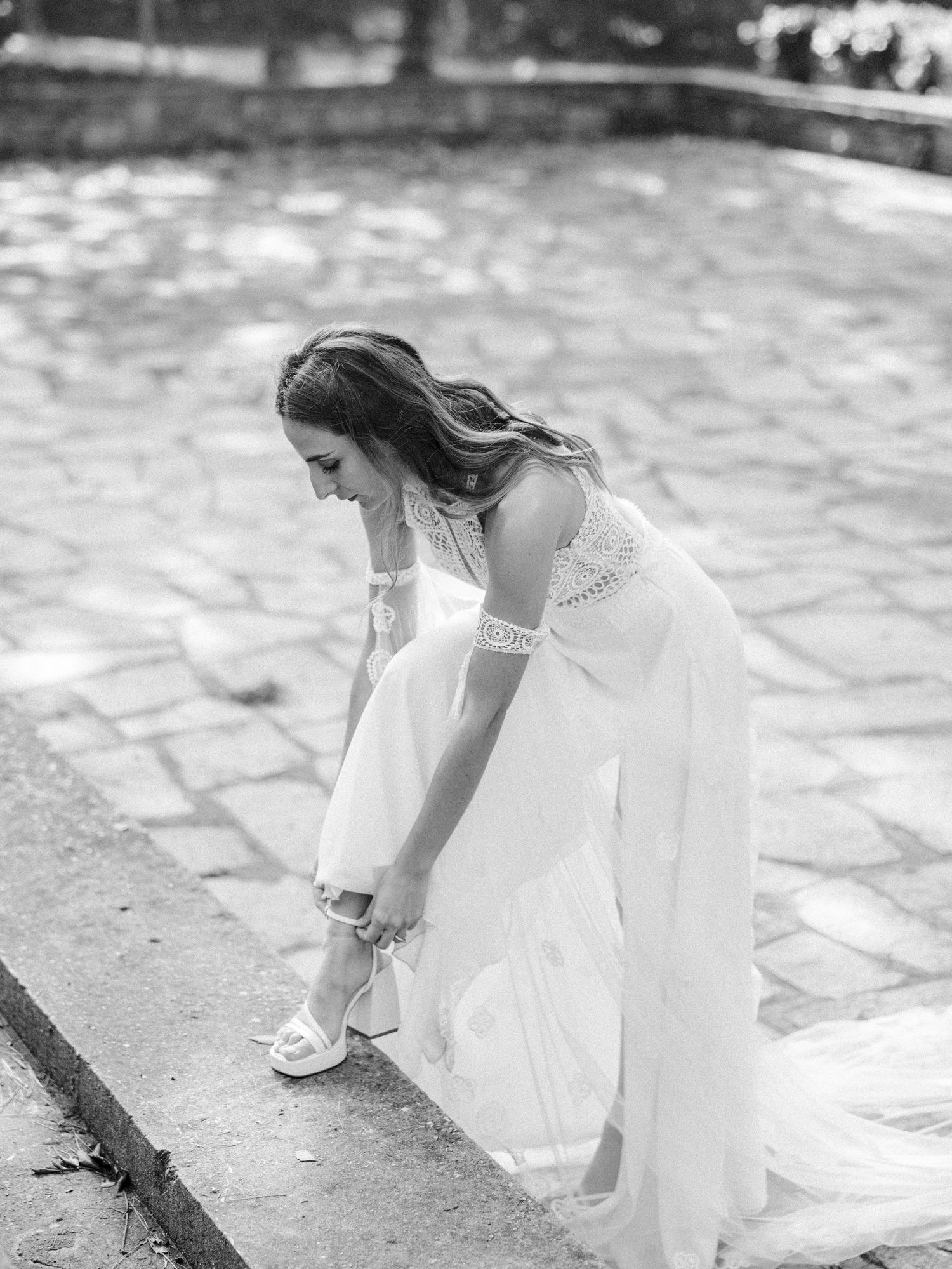 Black and white photo of bride adjusting her shoe on stone pathway.