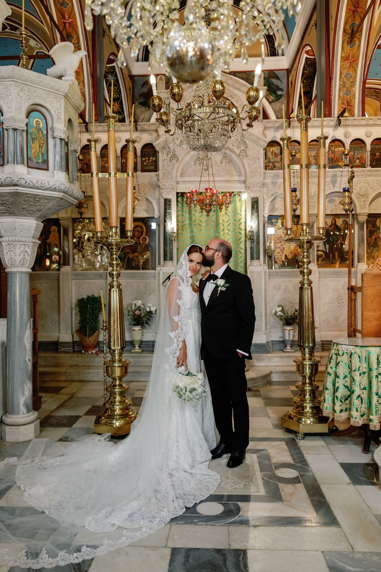 Bride and groom share a romantic moment inside an ornate Orthodox church with golden candelabras and chandelier.