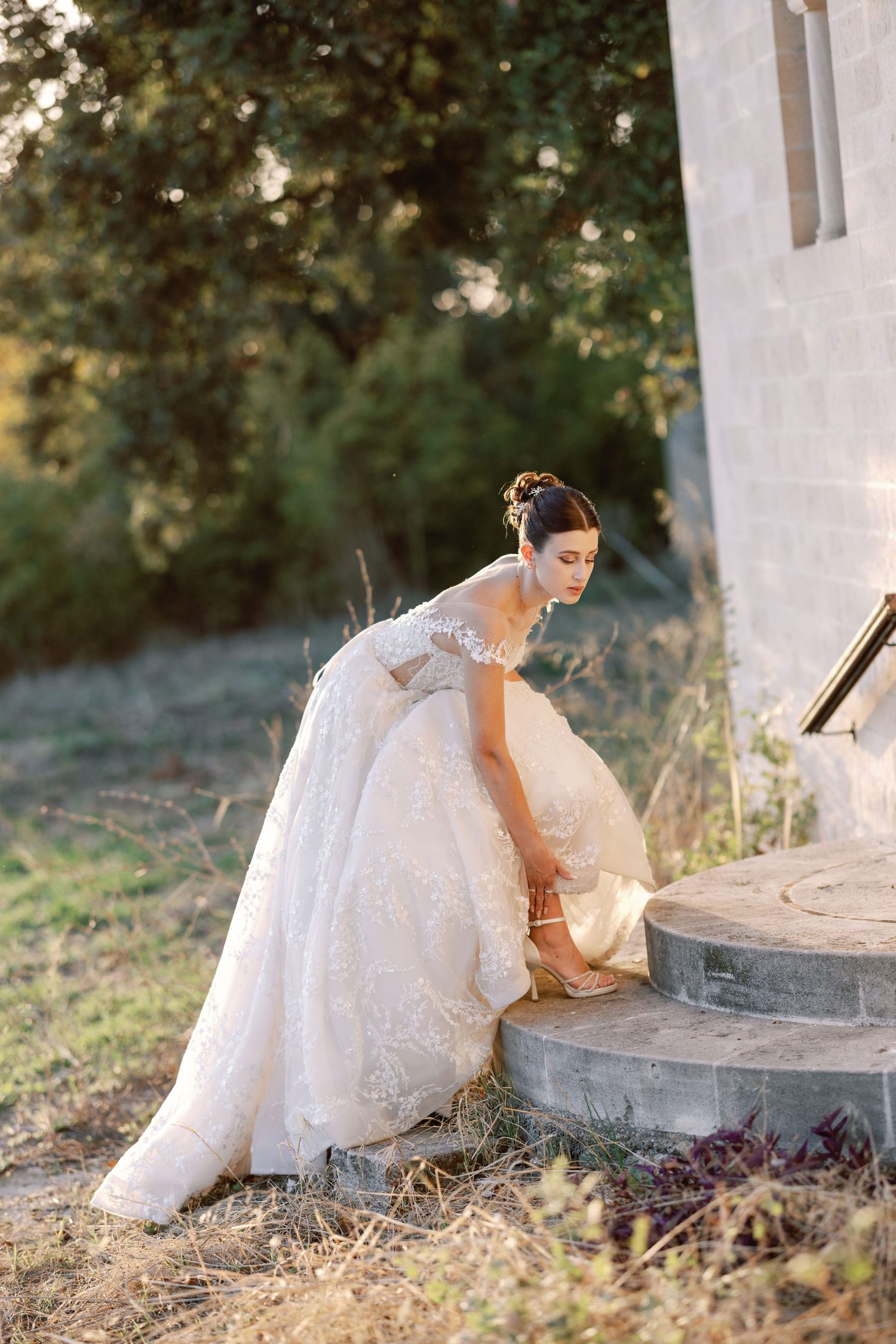 Bride in lace gown sits gracefully by white stone building in natural afternoon light.