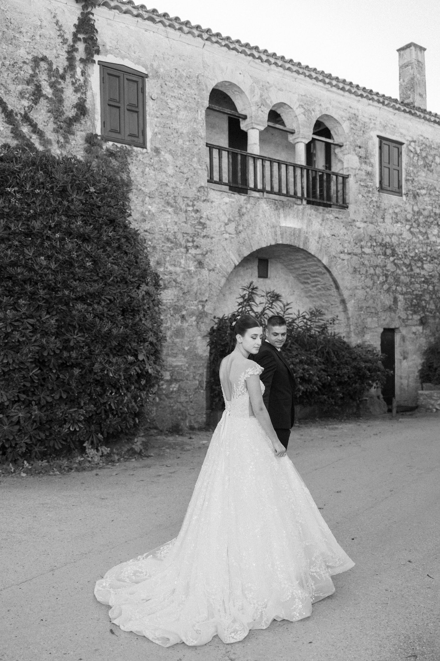 Elegant wedding couple walks together against stone building backdrop in romantic black and white photo series.