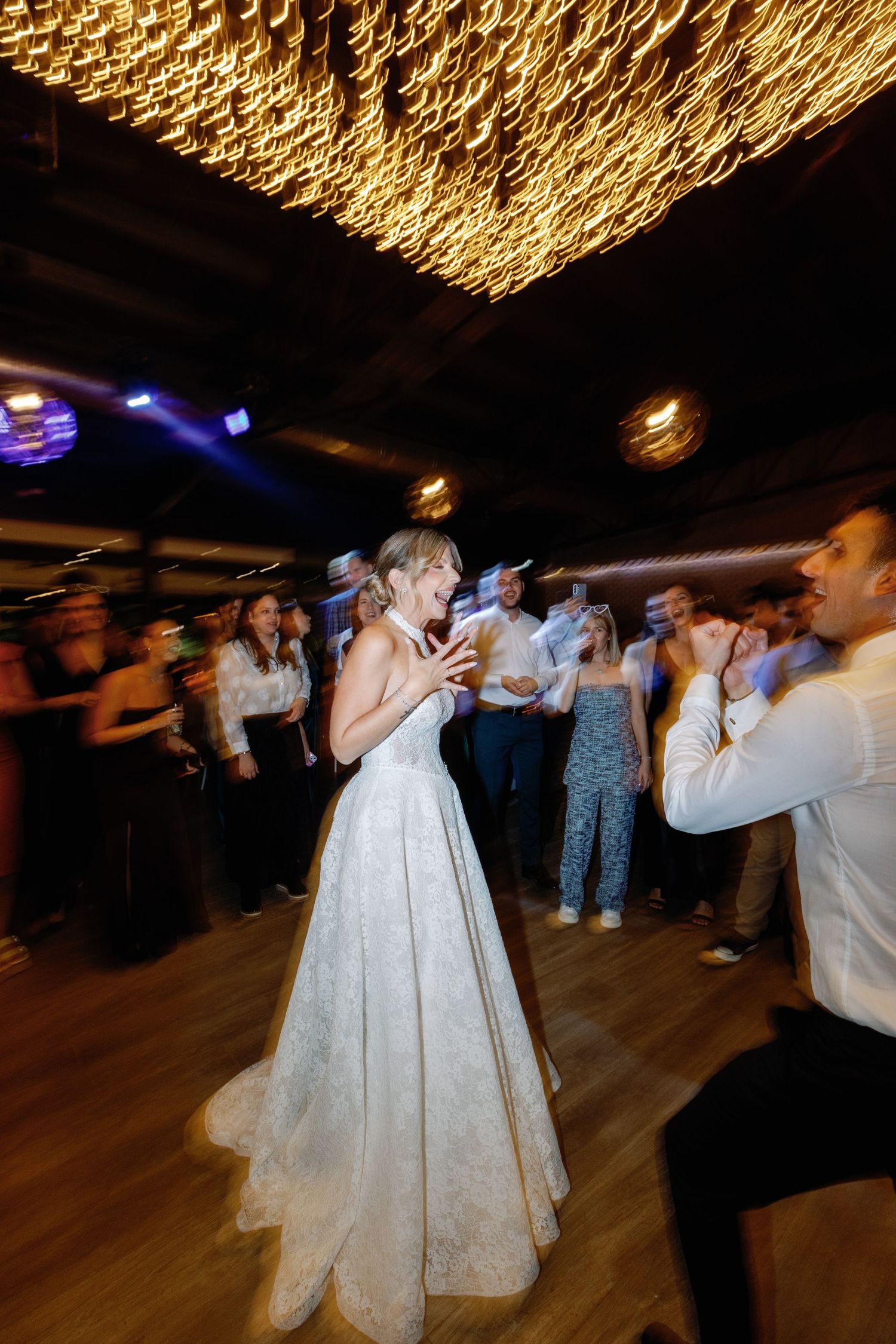Bride and guests dancing energetically under glowing ceiling lights at wedding reception.