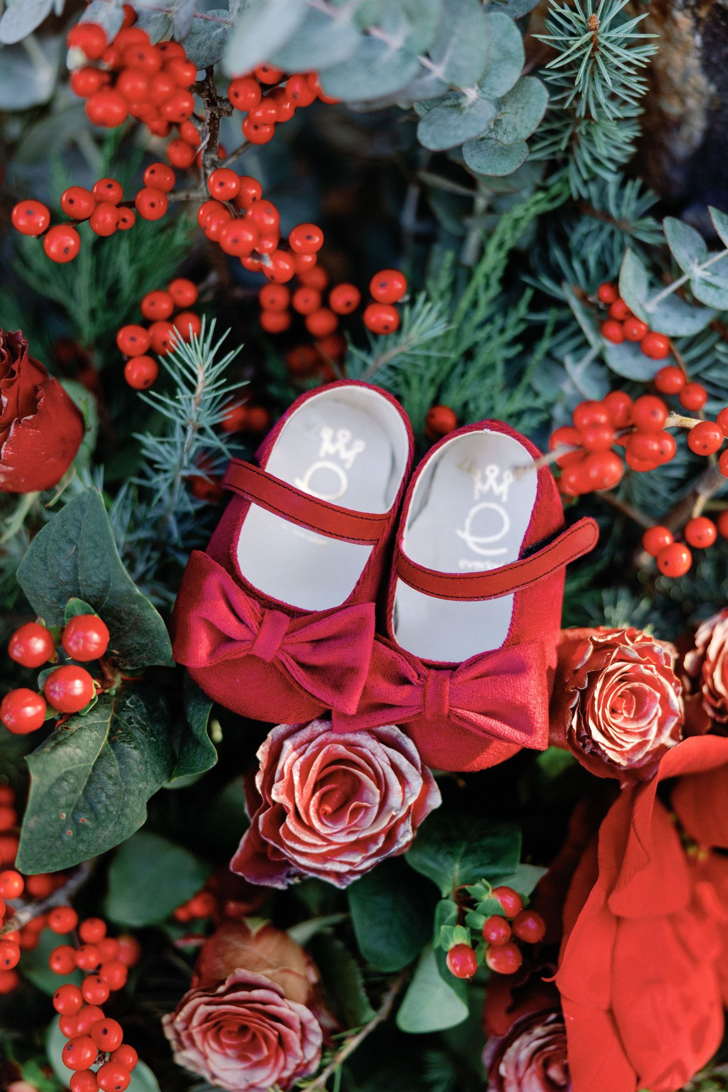 Bright red baby shoes with bows nestled among pink roses and festive red berries in a holiday floral arrangement.