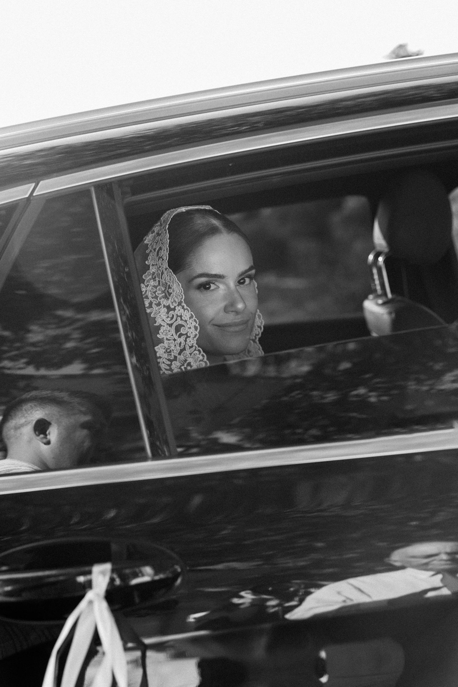 Black and white photo of a bride in lace veil sitting in a vintage car, gazing out the window on her wedding day.