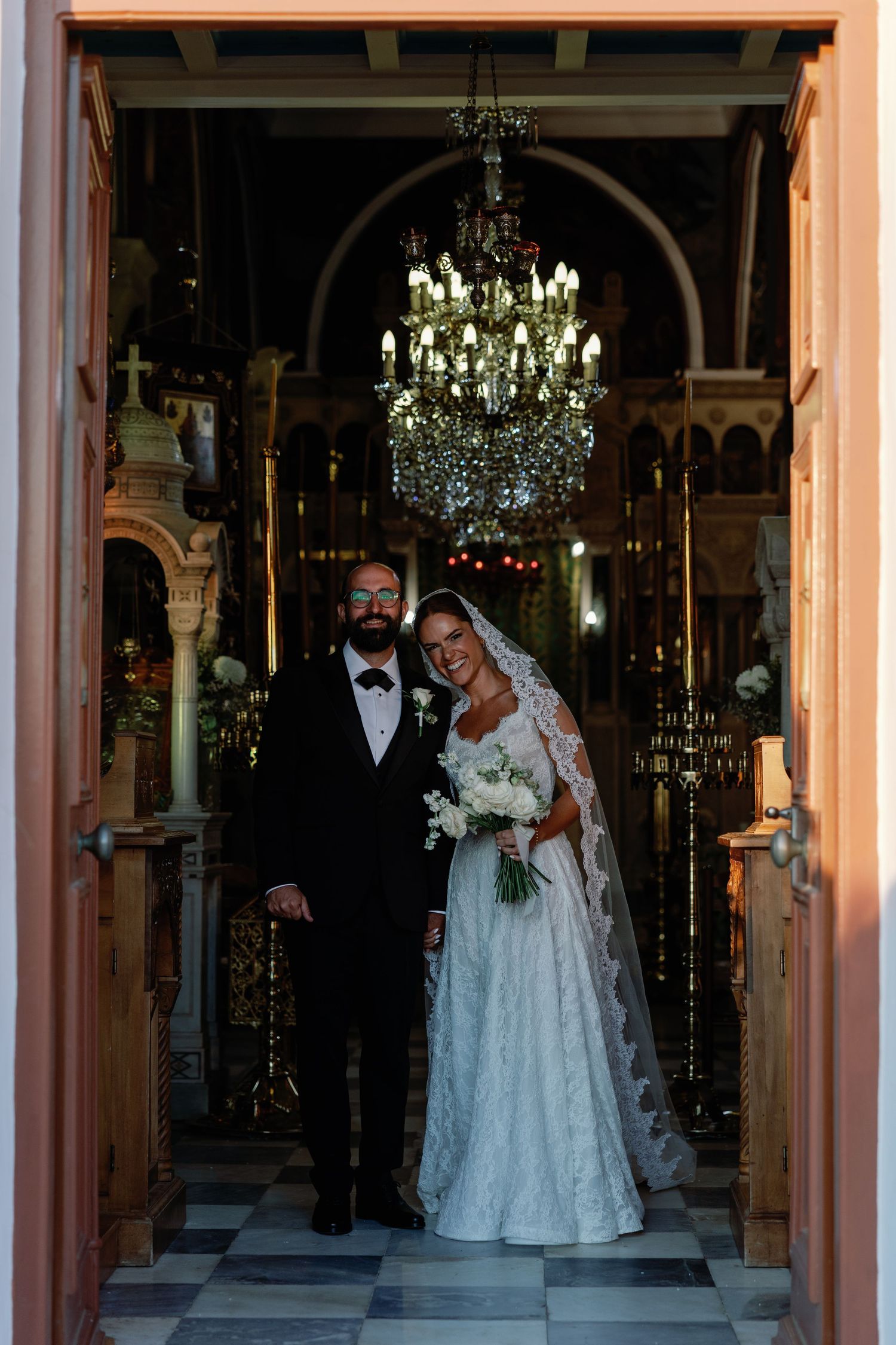 Bride and groom stand in church doorway under chandelier, she in lace gown with veil, he in black tuxedo.