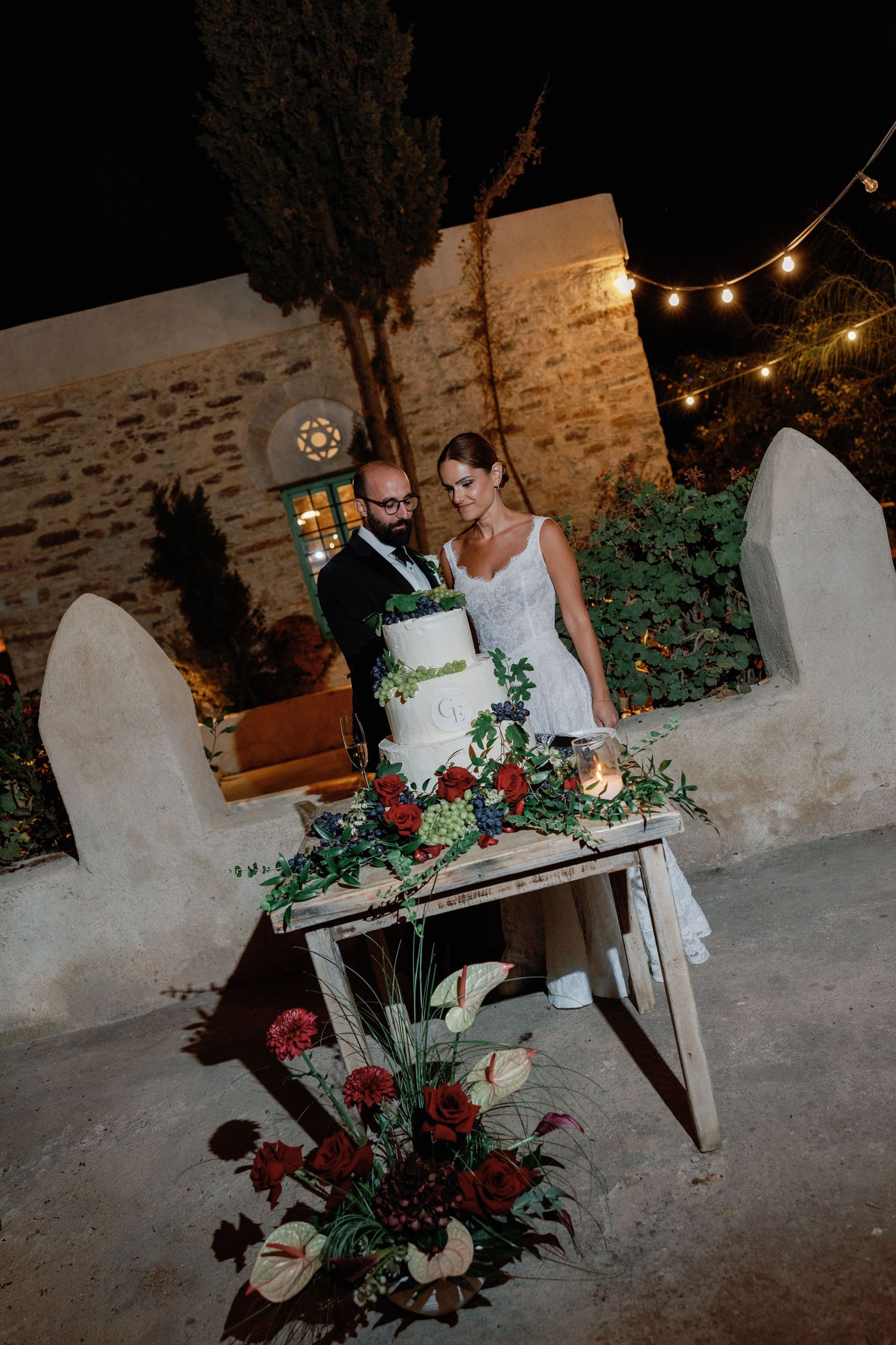Bride and groom cutting their wedding cake outdoors at night, surrounded by floral arrangements and string lights.
