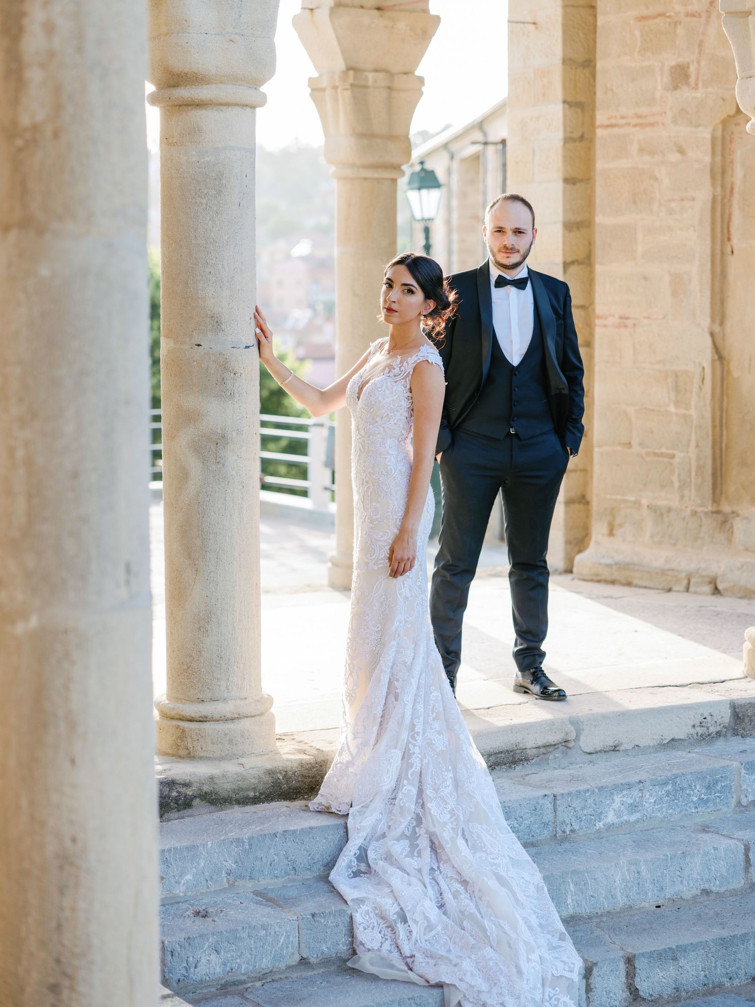 A couple in formal wedding attire poses beneath classical stone columns and arches.