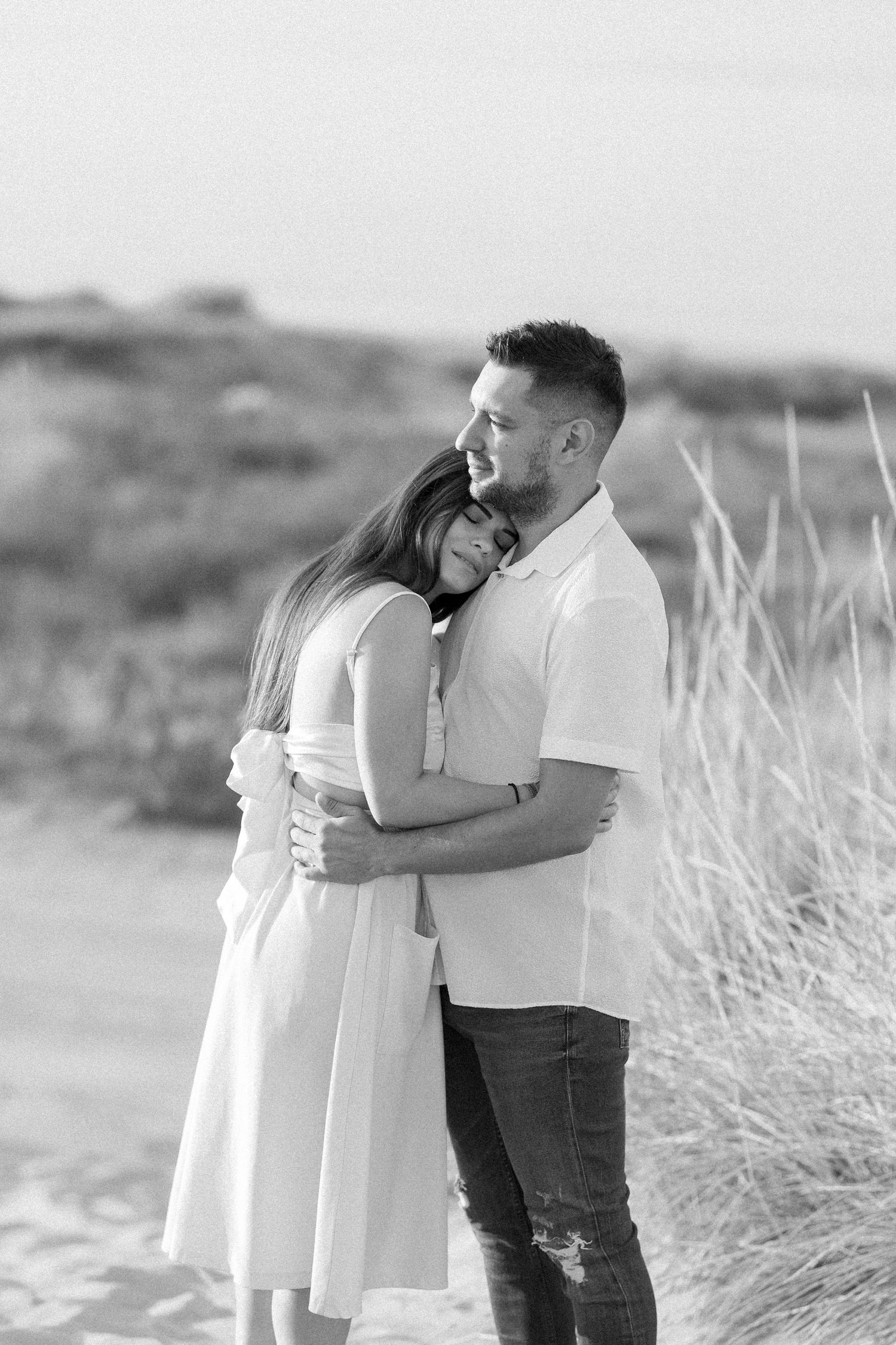 Couple embracing on a scenic beach with tall grass in black and white.