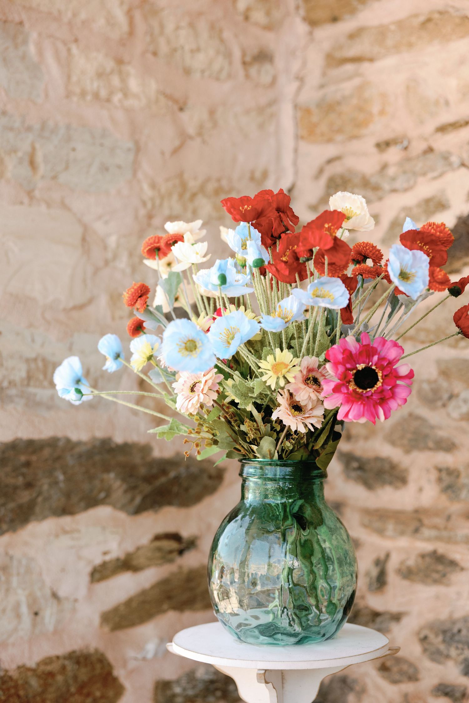 A colorful bouquet of flowers including poppies and daisies arranged in a blue-green glass vase against a textured stone wall.