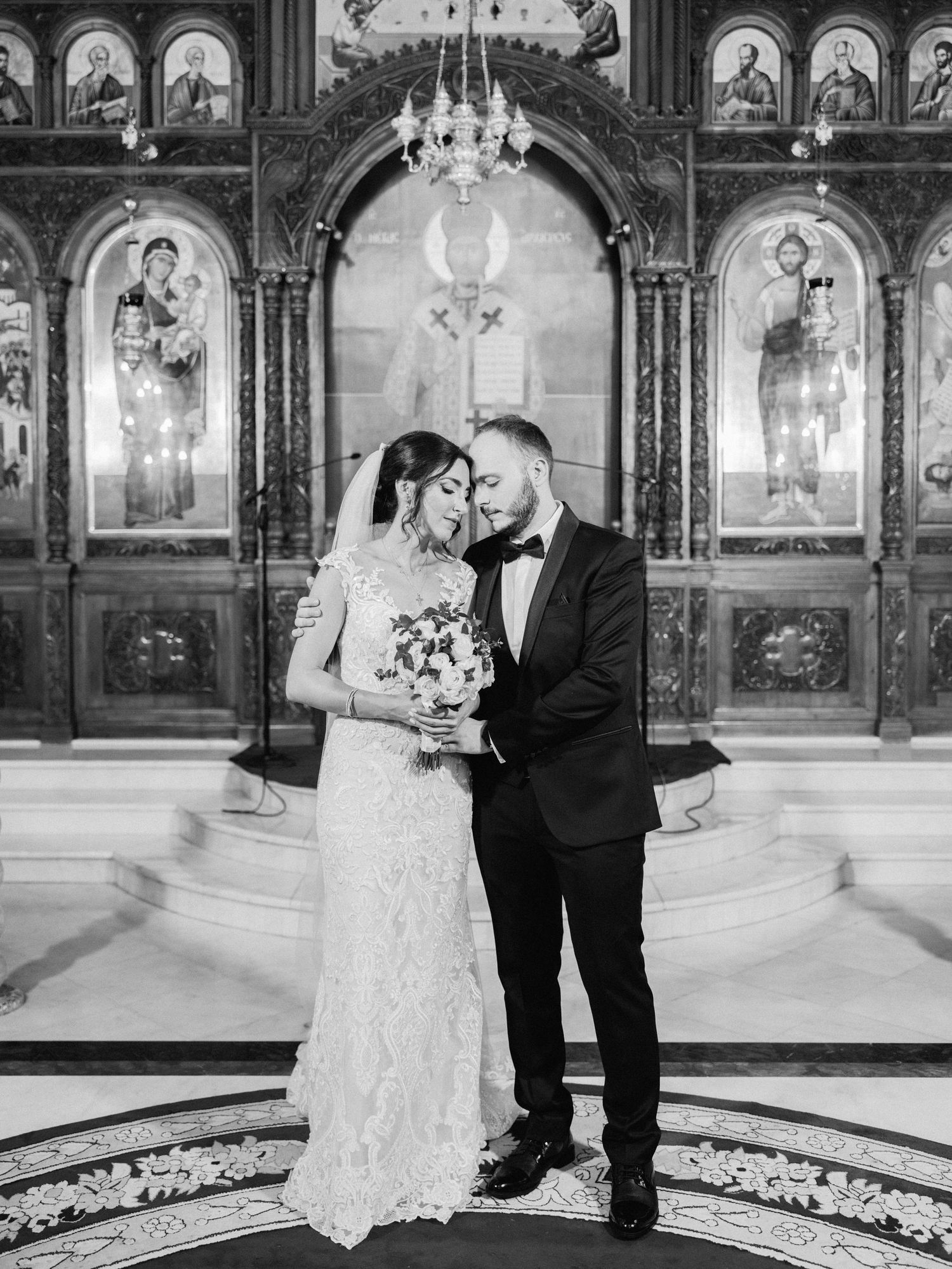 Elegant wedding couple embraces in front of ornate church iconostasis with chandelier and religious artwork.