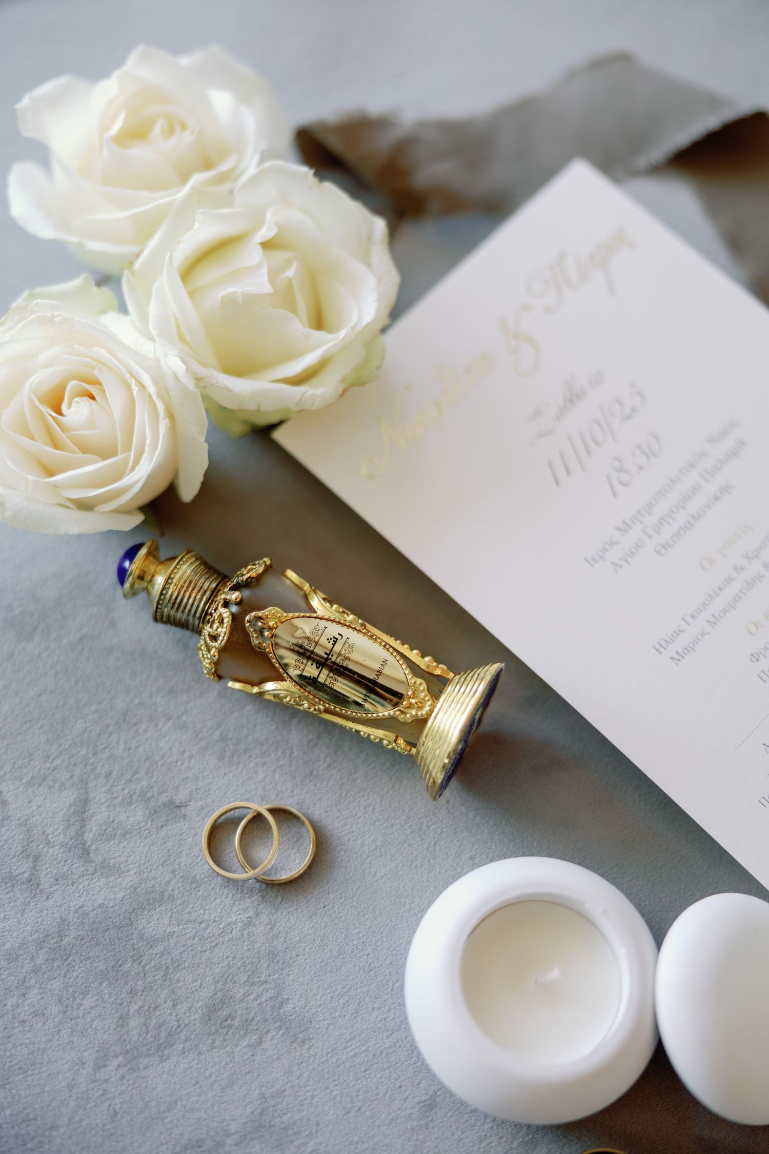 Wedding details flatlay with white roses, rings, invitation and decorative perfume bottle.