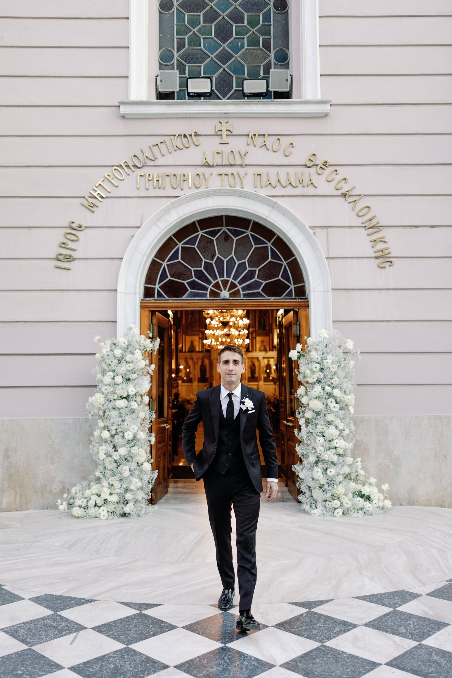 Groom in classic tuxedo walks through ornate church entrance decorated with white flowers.