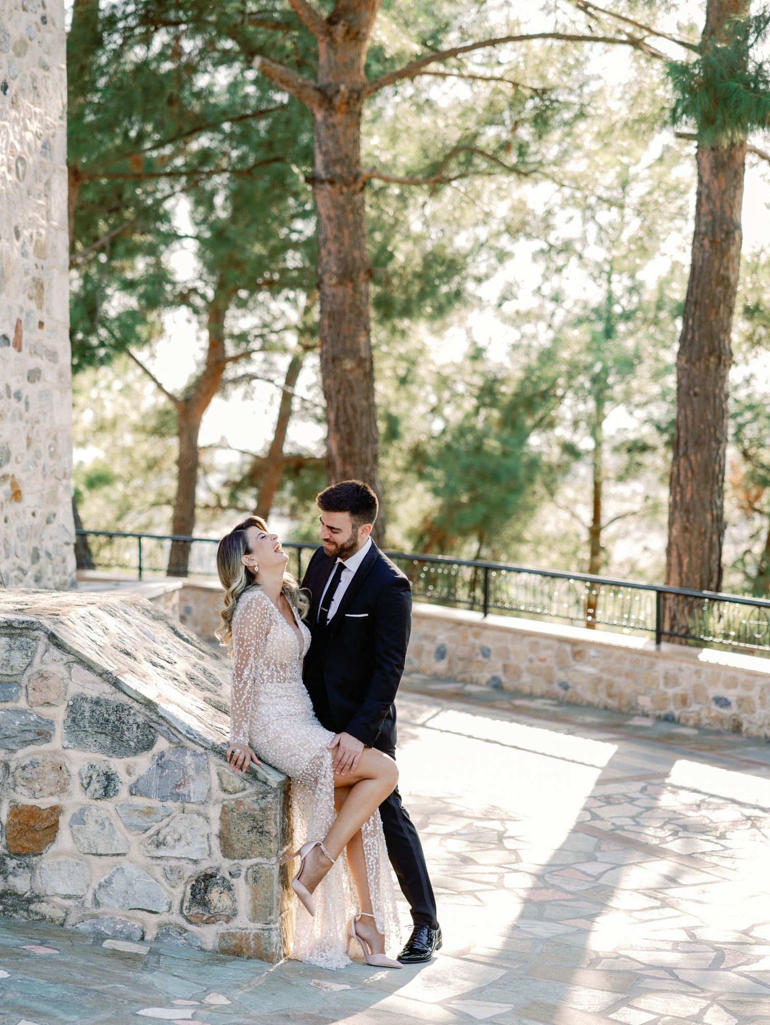 Playful wedding couple shares romantic moments on stone patio with pine trees in background.