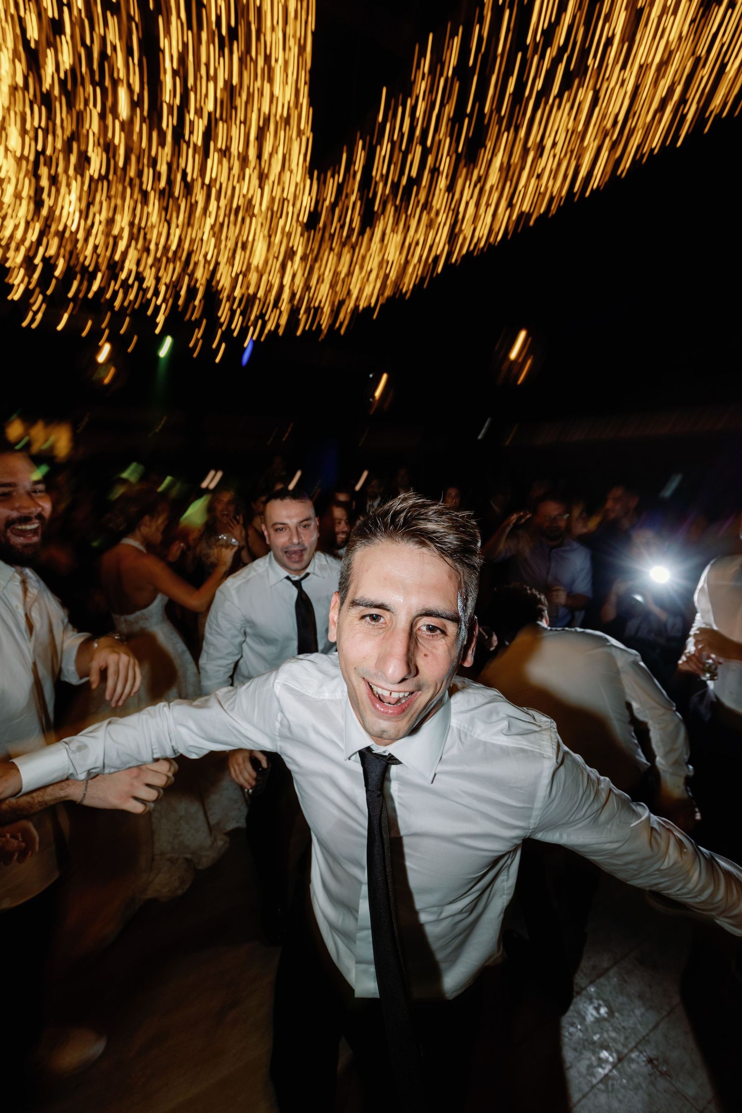 Wedding guests dancing energetically under golden string lights at reception.