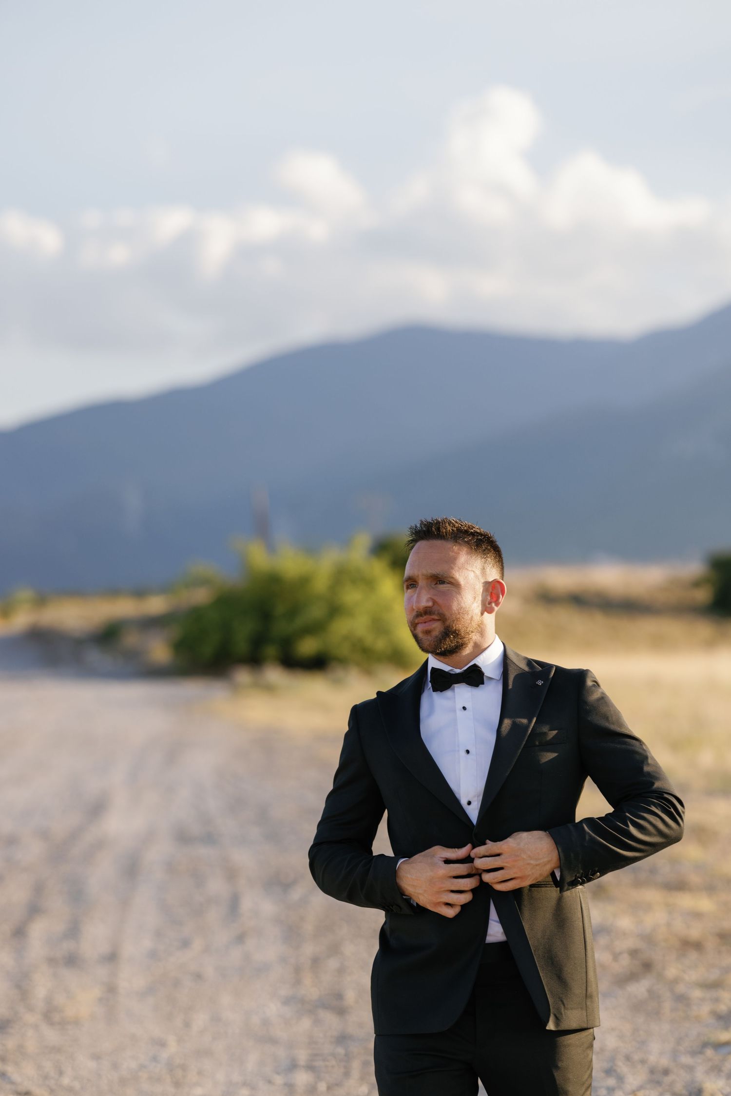 A groom in a black tuxedo poses against a mountainous backdrop during a wedding photoshoot.