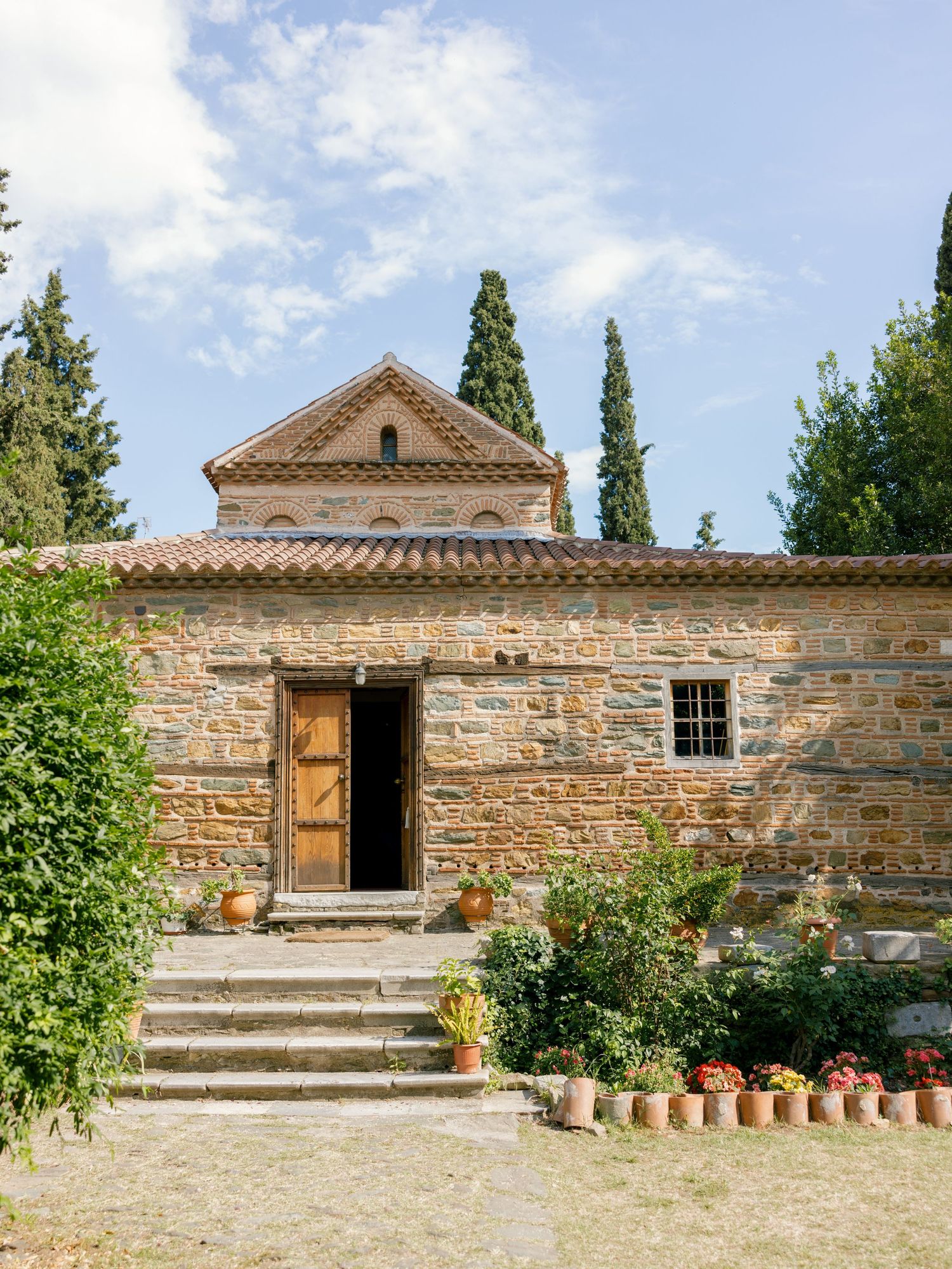 Ancient stone church with terracotta roof tiles, wooden door, and potted plants along stone steps in Mediterranean setting.
