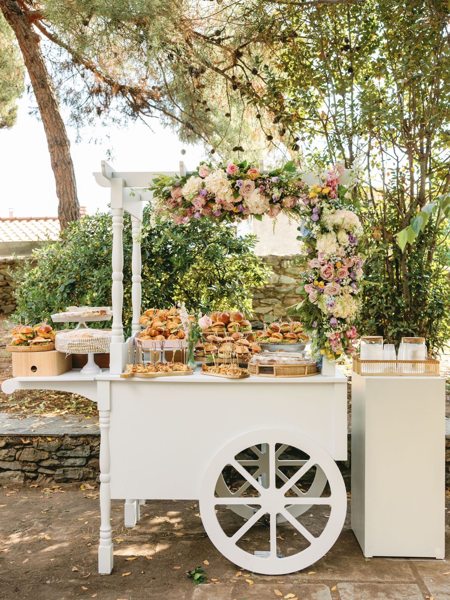 A rustic white wooden cart decorated with floral arrangements serves as an elegant outdoor display piece at a garden event.