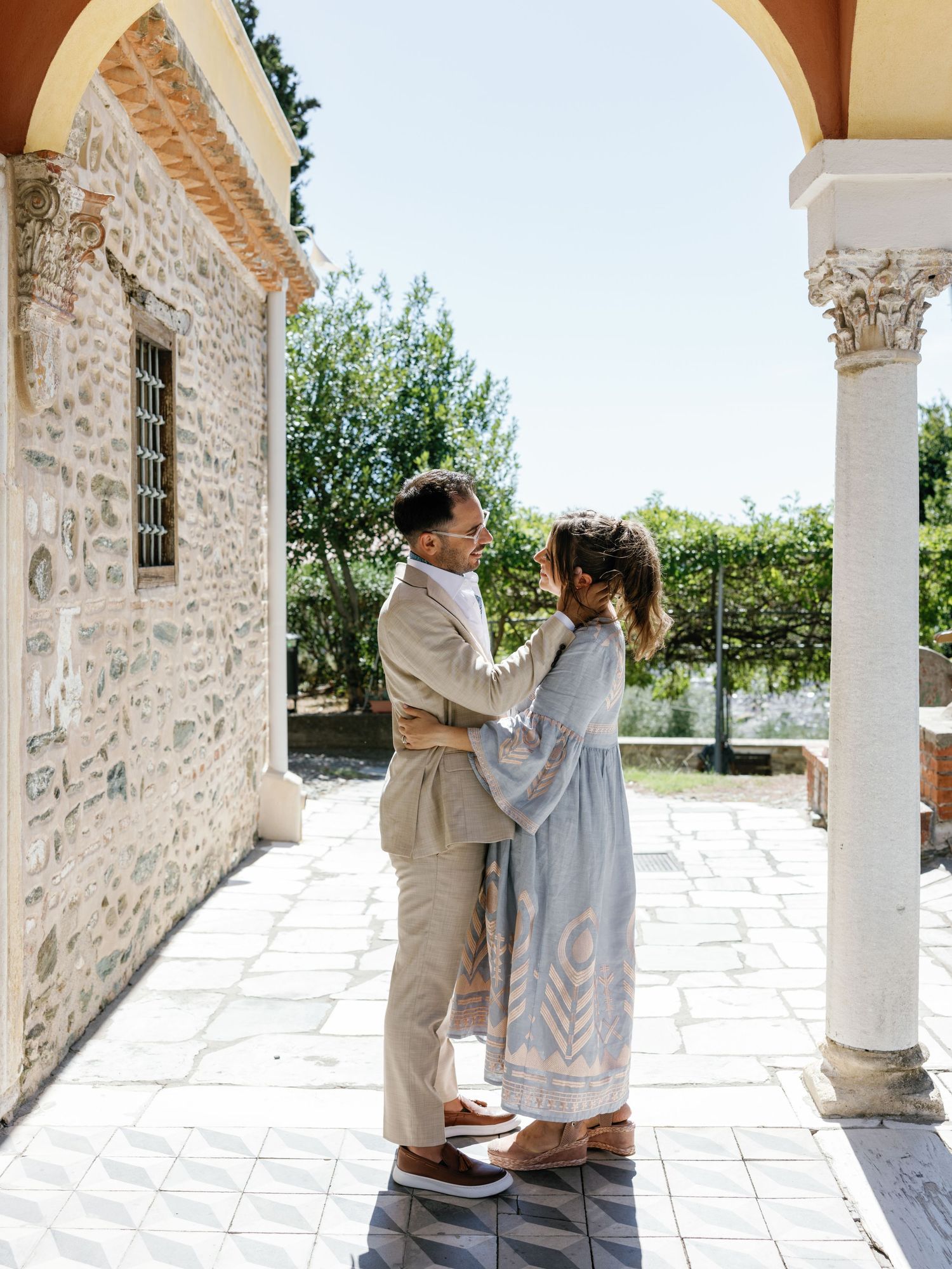 A couple in light colored clothing embraces on a Mediterranean style porch with stone walls and white columns.