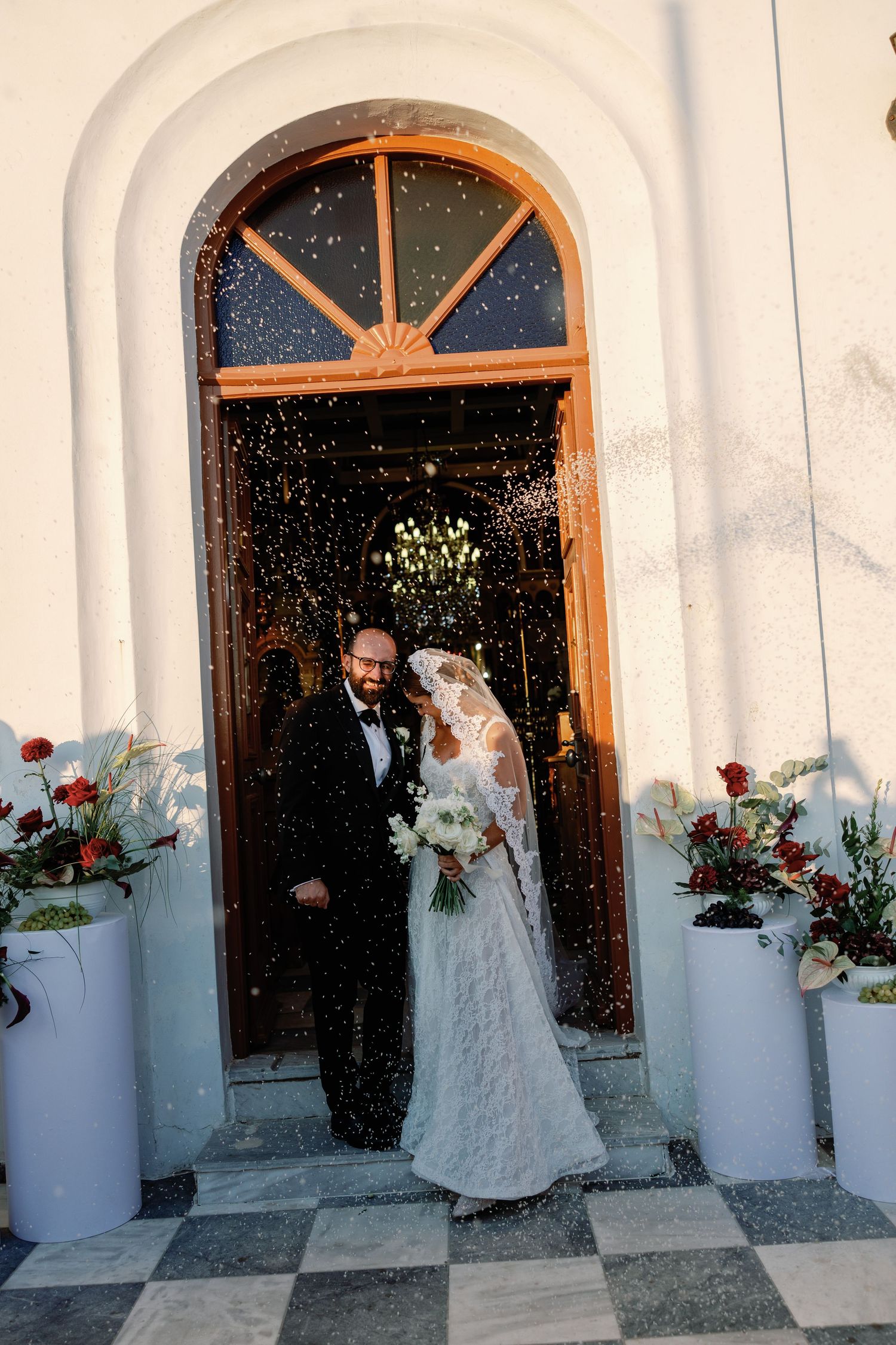 Newlywed couple exits church doors in confetti shower, bride in lace gown with veil, groom in black tuxedo.