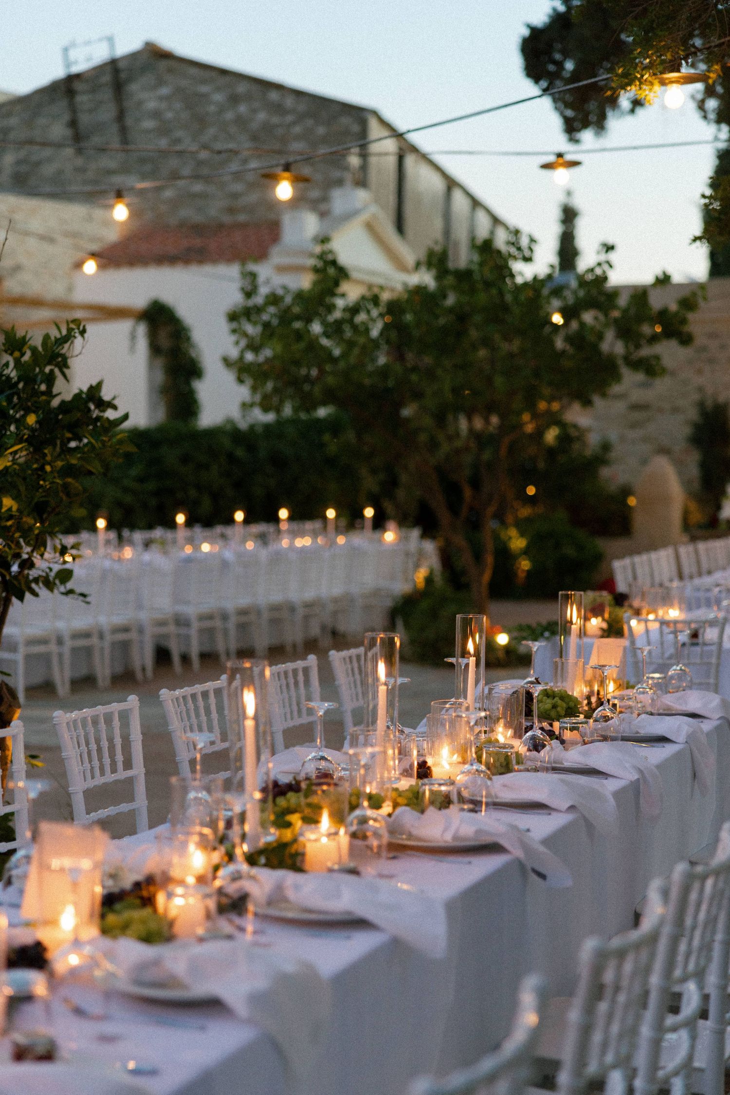 Elegant outdoor wedding reception with long white tables, candlelight, string lights, and lush greenery at dusk.